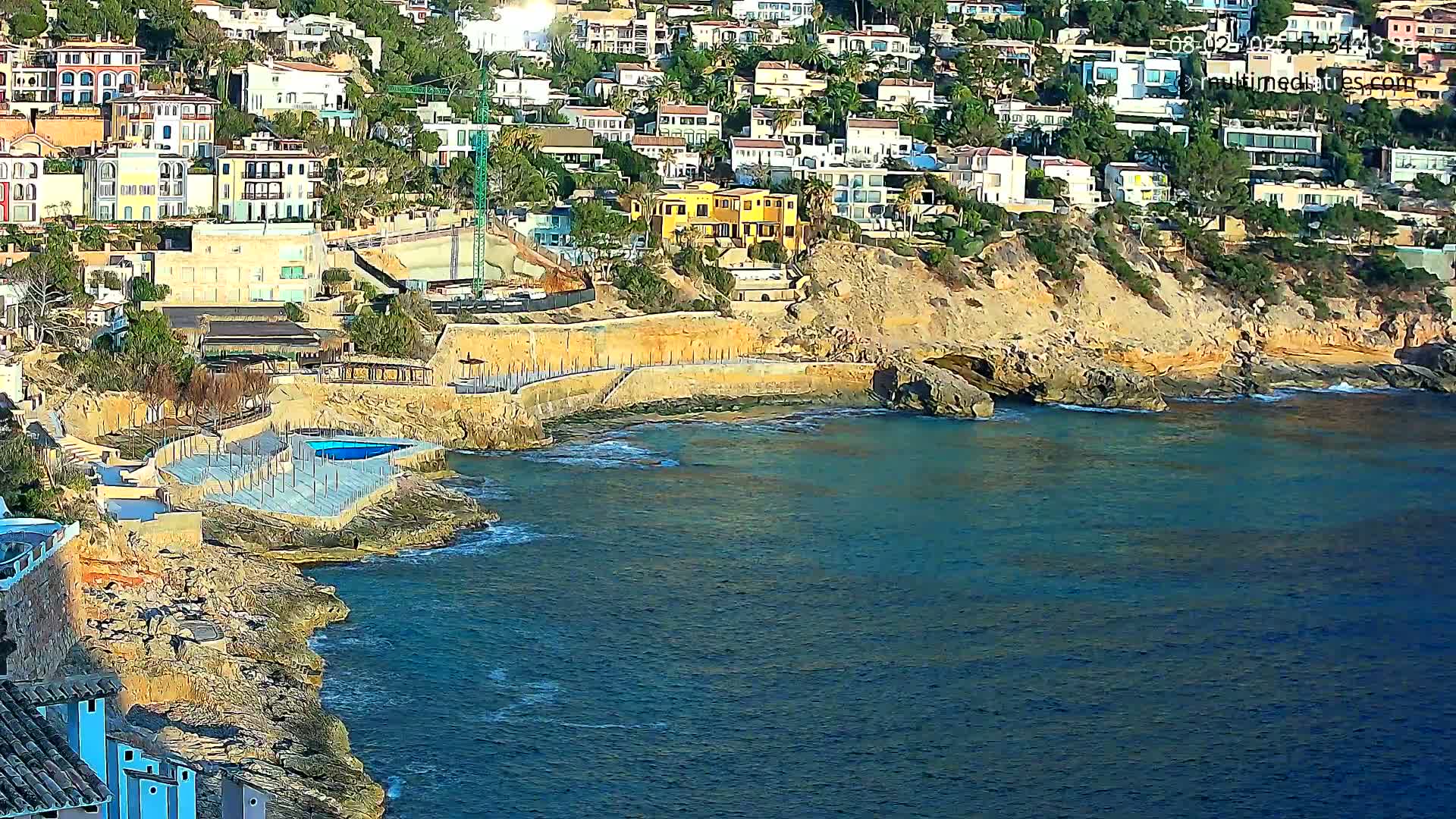 A rocky coastline with a small swimming pool and numerous houses on a hillside overlooking a calm, blue sea.