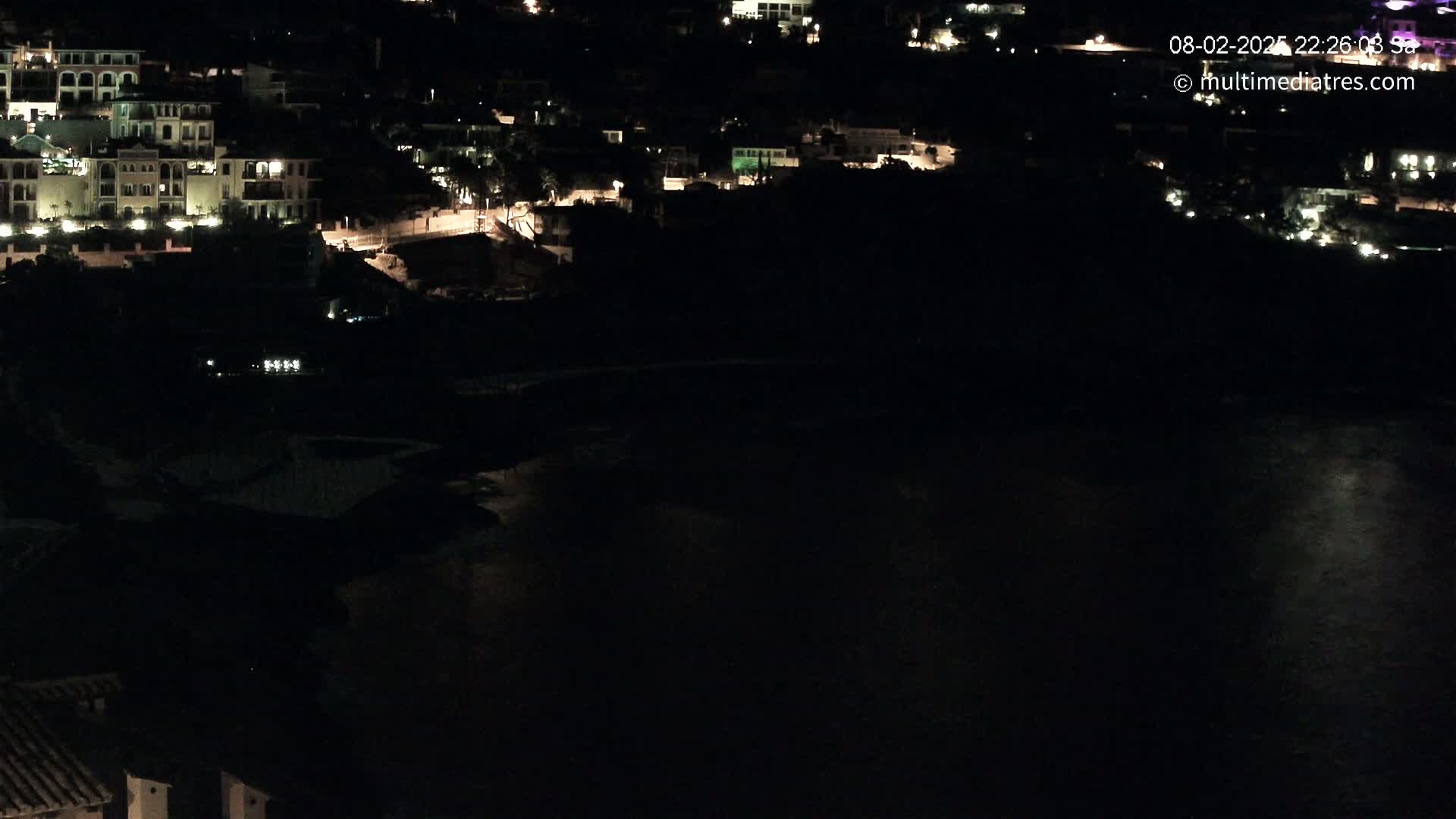 A nighttime view shows a dark body of water below a hillside of dimly lit buildings.