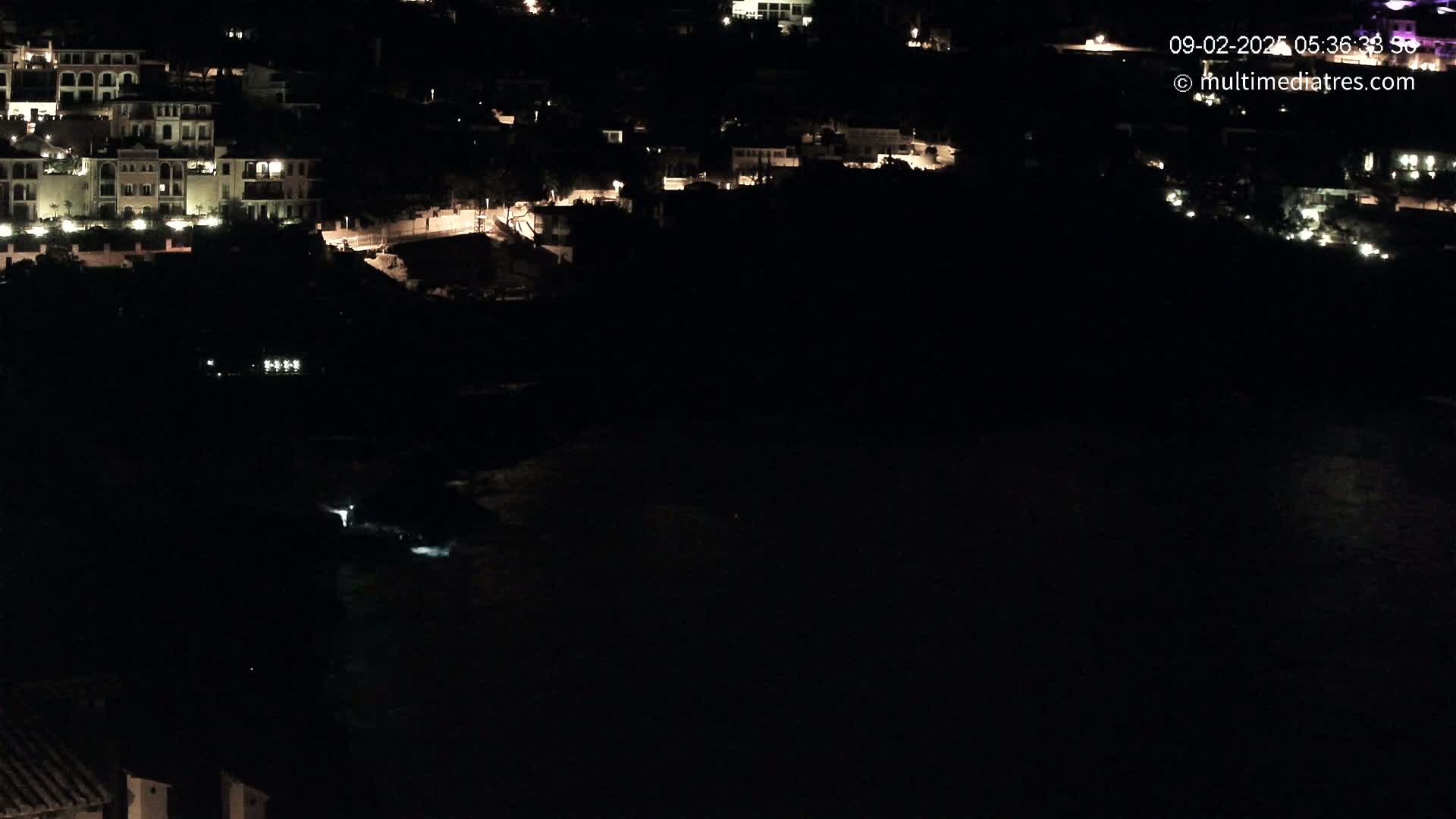 A nighttime, high-angle view shows a dark body of water below a coastal town with dimly lit buildings.