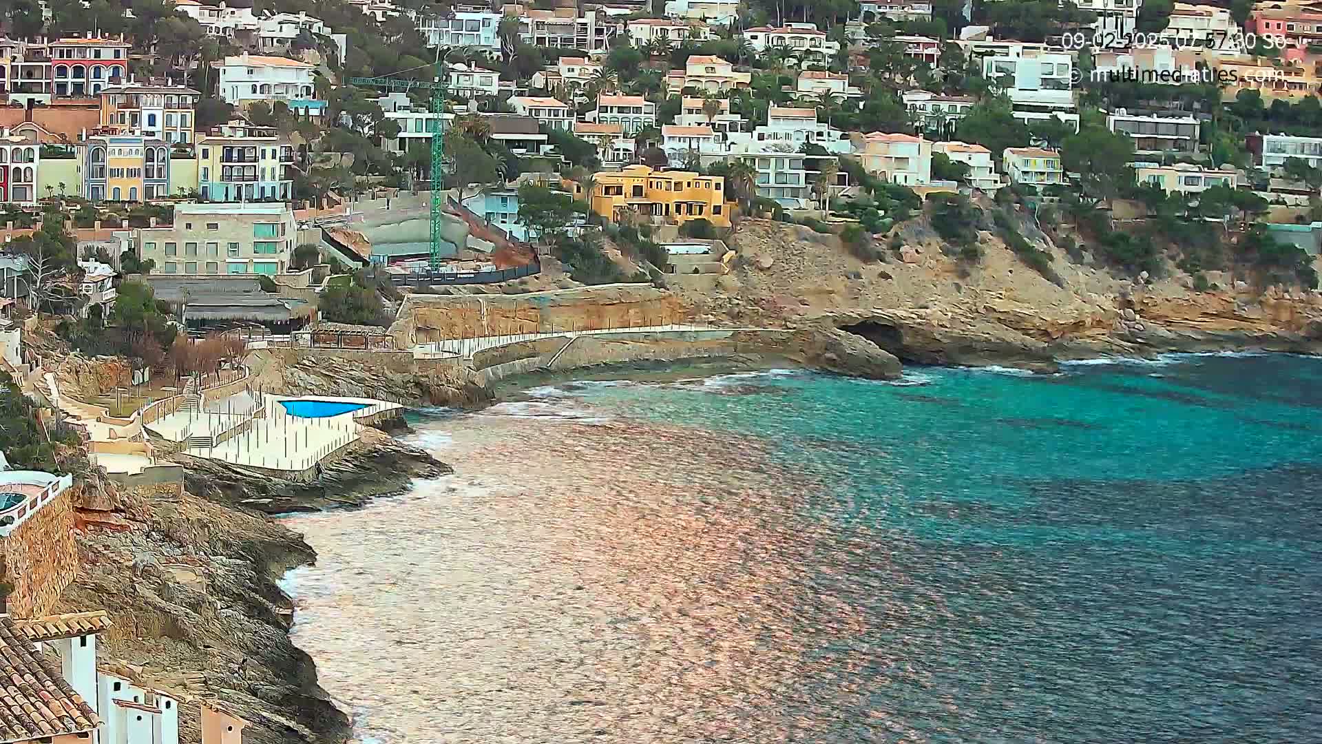 A rocky coastline with turquoise water features a small beach, a swimming pool, and a hillside of houses.
