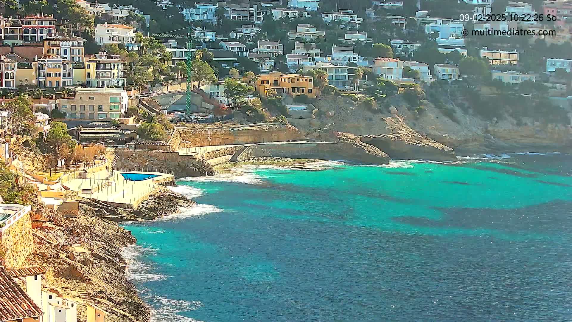 A rocky coastline with turquoise water features a small swimming pool and many houses built into the hillside on a sunny day.