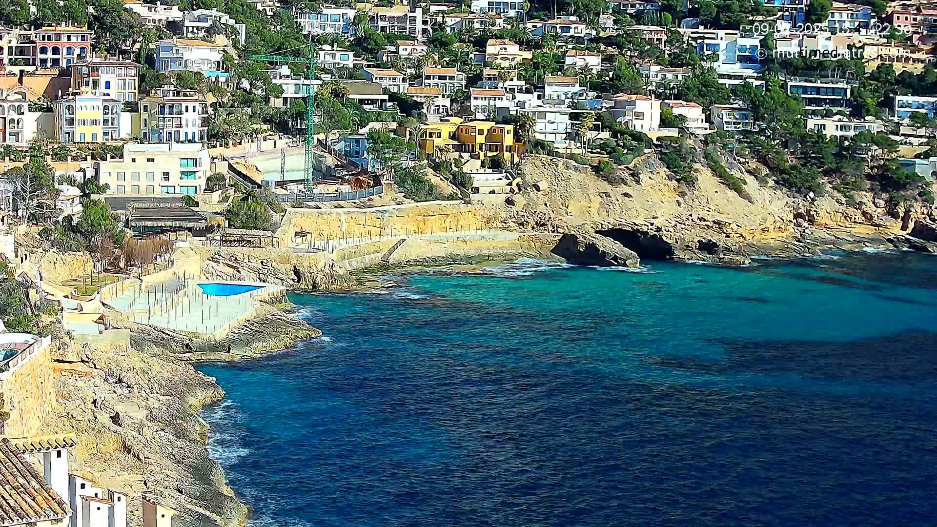 A rocky coastline with turquoise water features many houses built into a hillside, and a small swimming pool is visible near the water's edge.