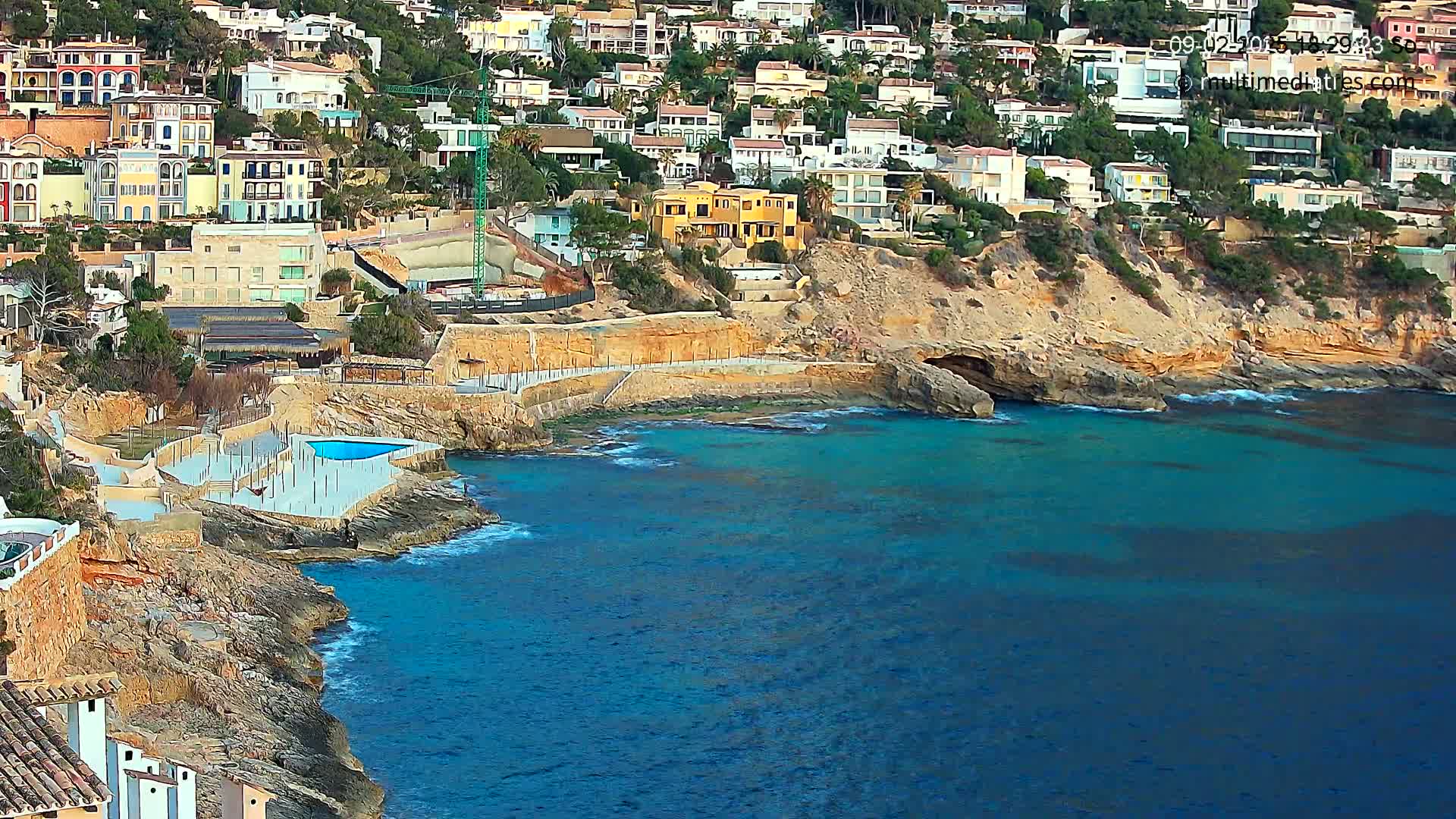 A rocky coastline with turquoise water features a cluster of buildings on a hillside, a small pool, and some light wave action.