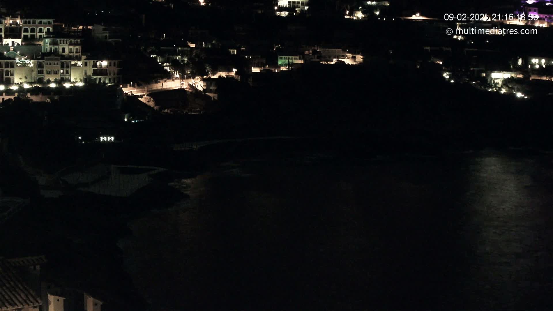 A nighttime, aerial view shows a dark body of water reflecting some light, with a hillside of illuminated buildings behind it.