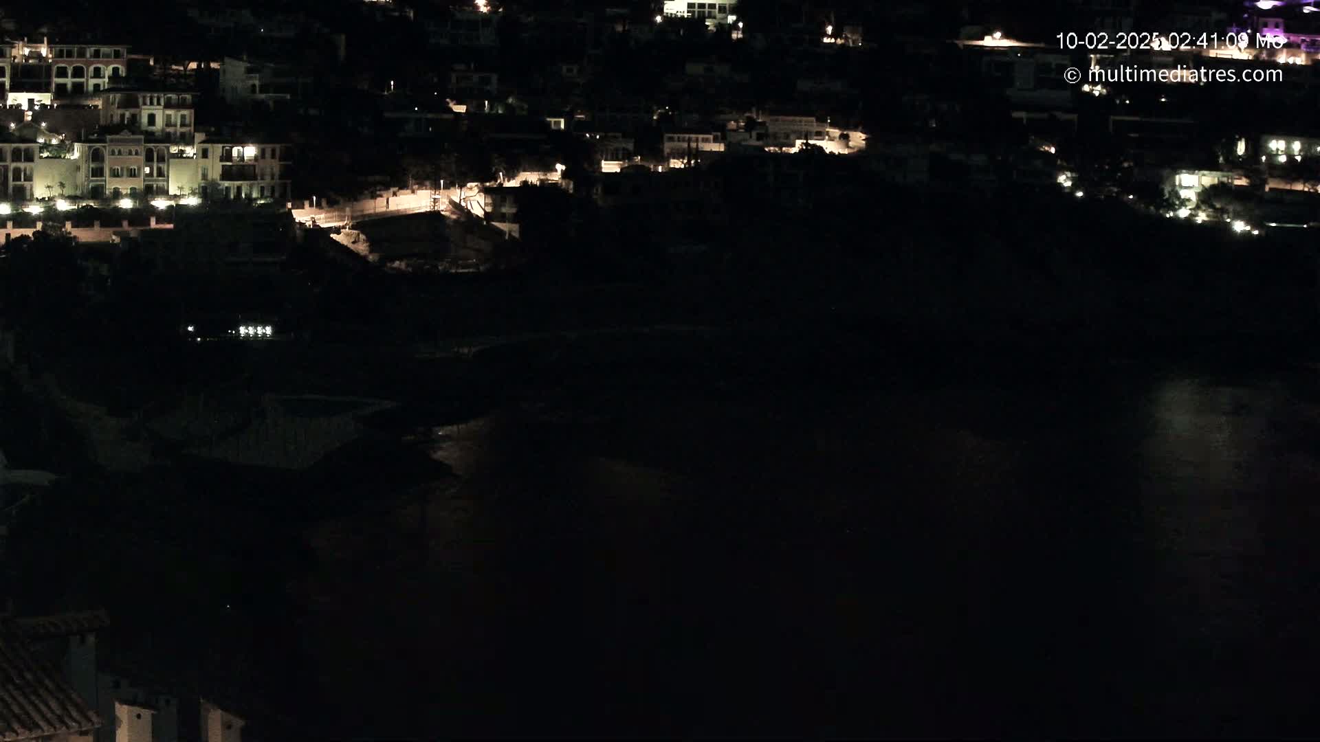 A nighttime view of a coastal town with brightly lit buildings overlooking a dark body of water.