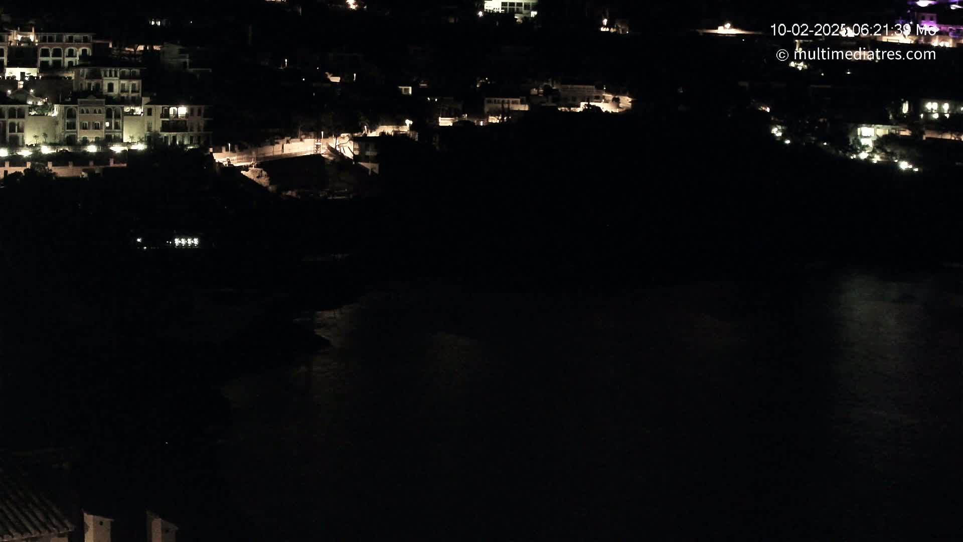 A nighttime aerial view shows a dark body of water below a hillside of illuminated buildings.