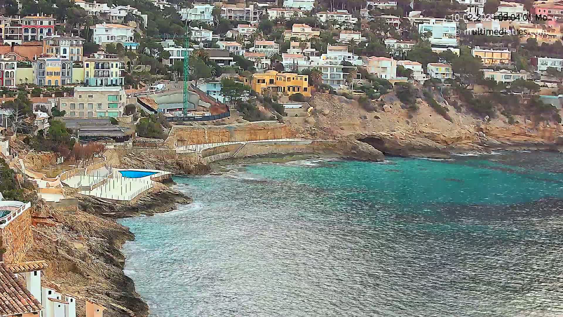 A rocky coastline with turquoise water features numerous houses built into a hillside, a small swimming pool, and some construction activity.