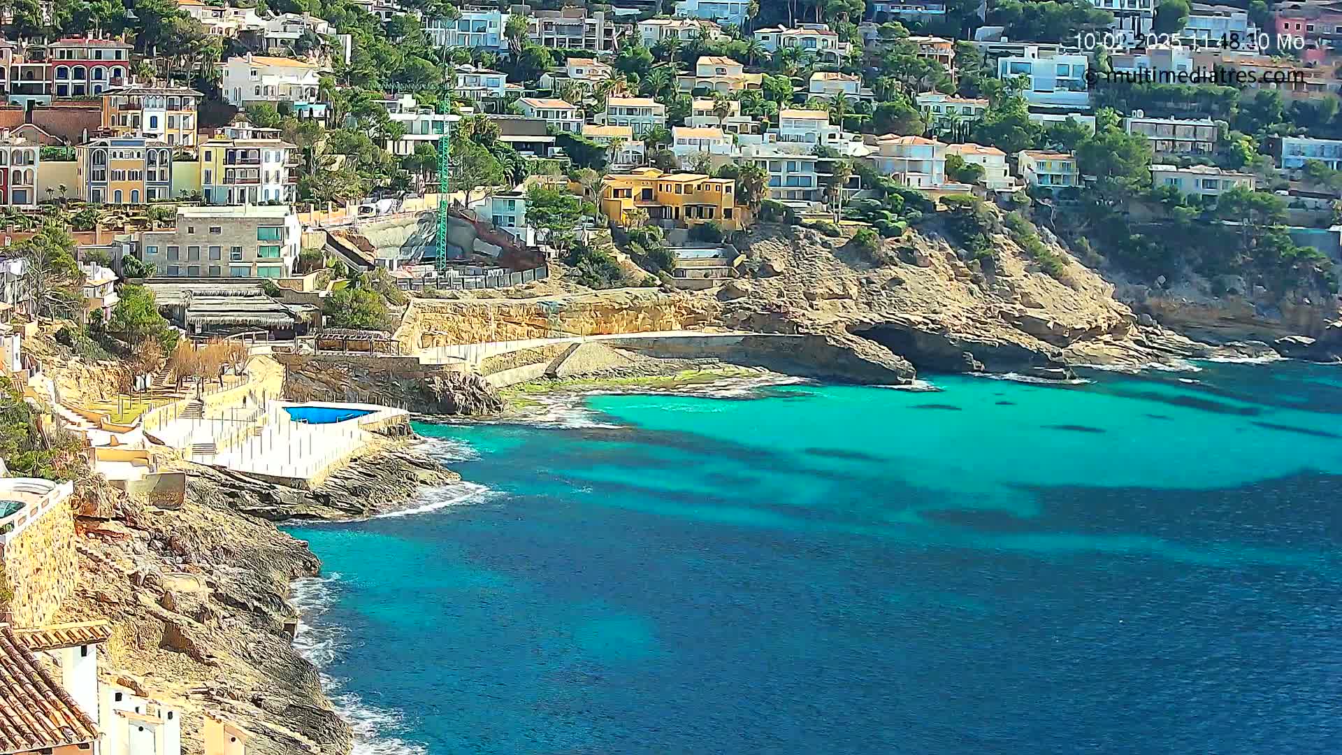 A hillside of houses overlooks a rocky cove with vibrant turquoise water on a sunny day.