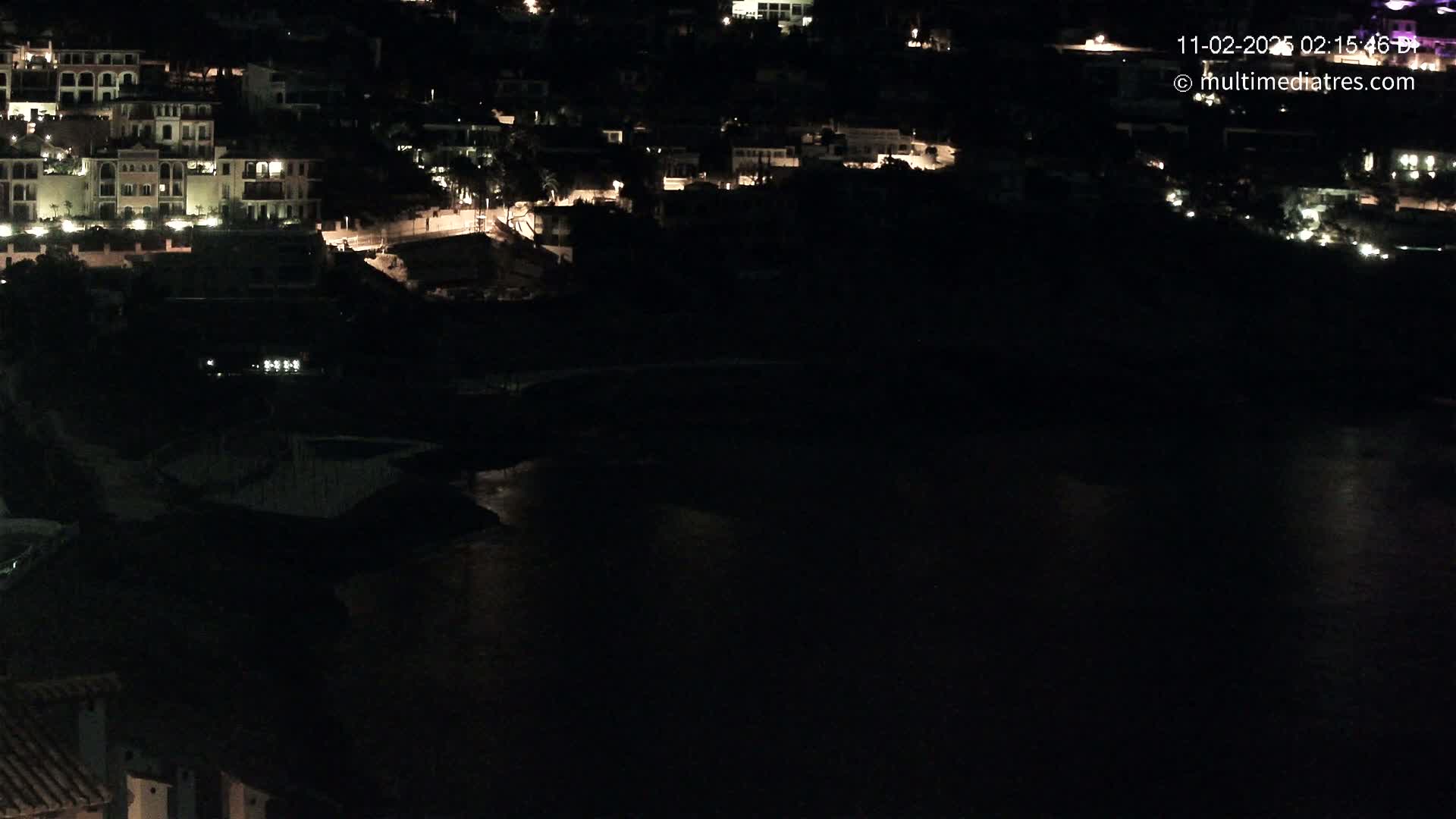 At night, a collection of buildings with illuminated windows sits on a dark hillside overlooking a calm body of water.