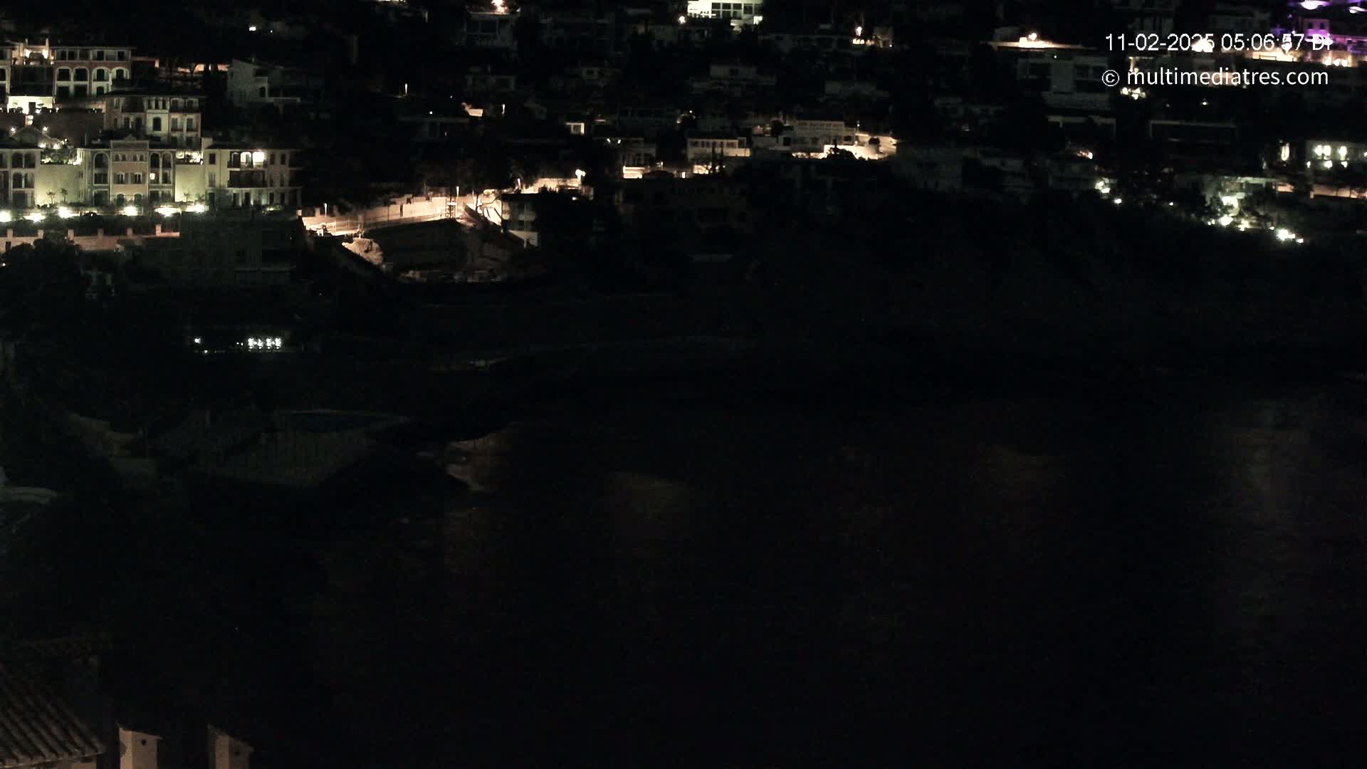 A nighttime, high-angle view shows a dark body of water reflecting the lights of a hillside town.