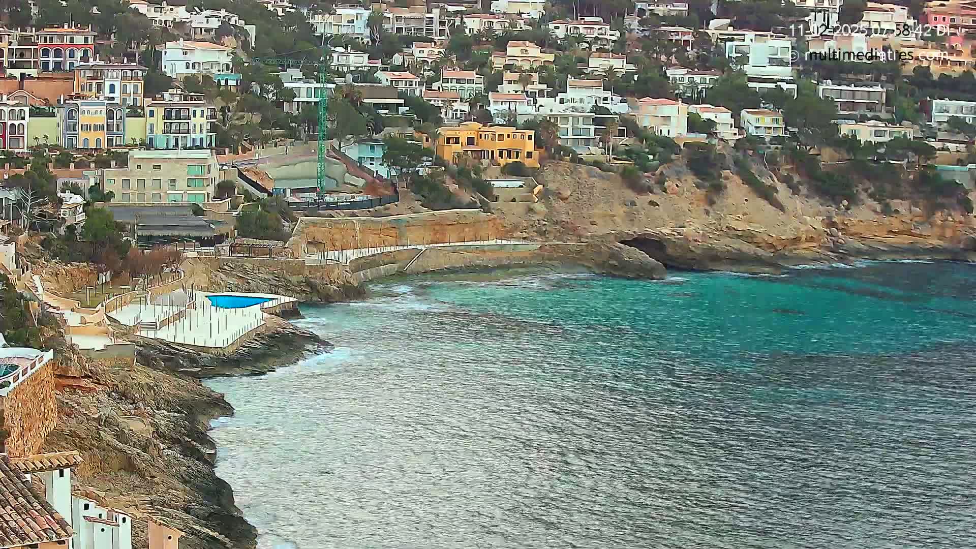 A coastal scene shows a collection of houses built into a hillside overlooking a calm, teal sea with a small pool situated on the rocky shoreline.