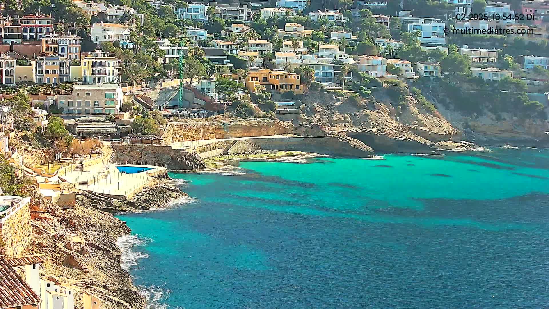 A rocky coastline with turquoise water, featuring a small swimming pool and numerous houses built into a hillside on a sunny day.