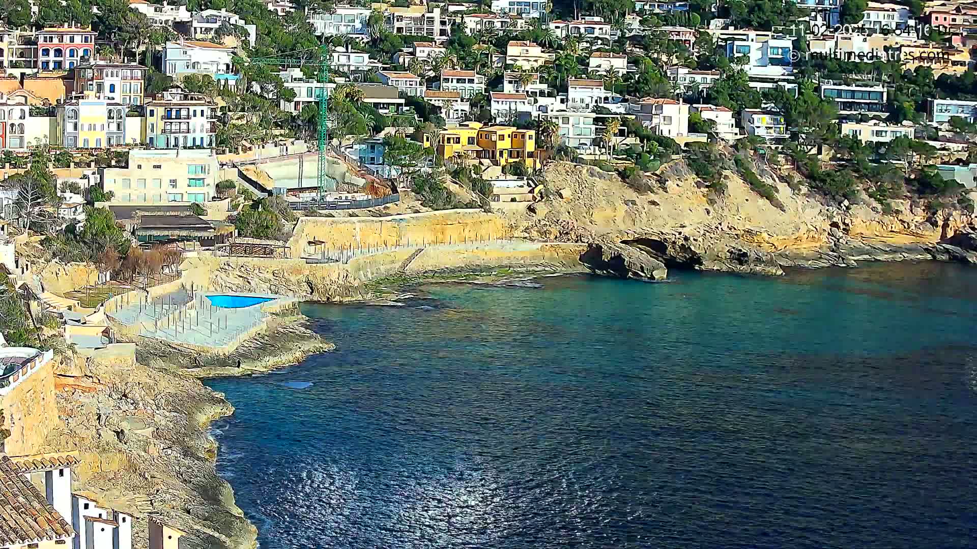 A coastal town with many houses built on a cliff overlooking a calm, teal-colored sea on a sunny day.