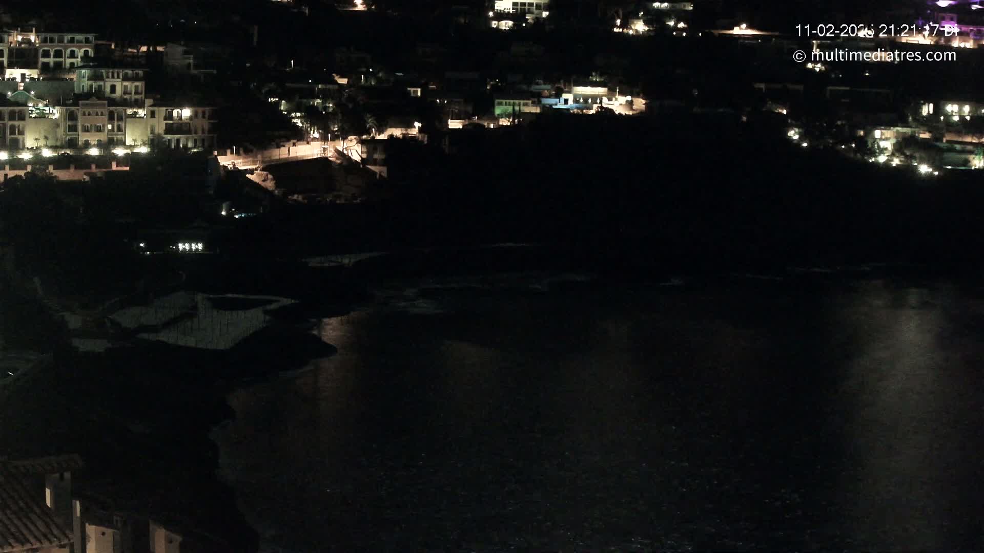 A nighttime view of a dark body of water reflecting lights from buildings on a hillside.