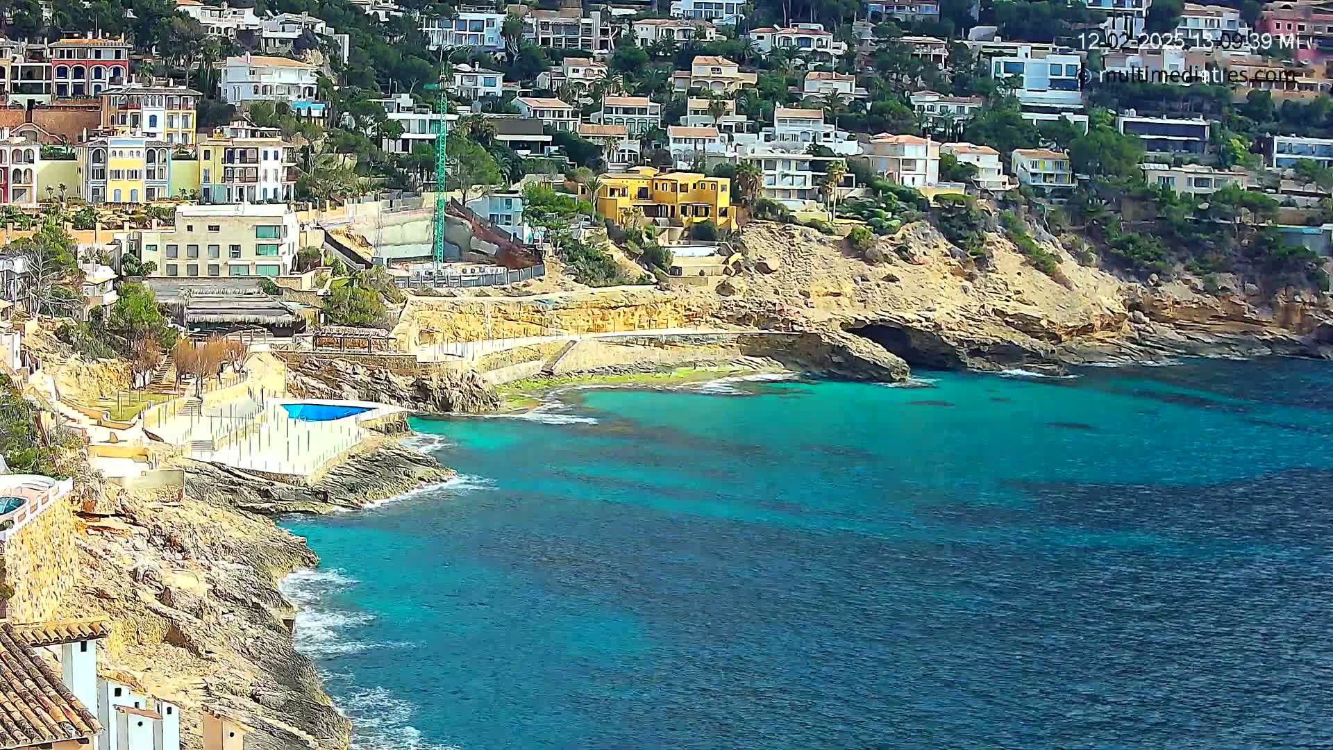 A rocky coastline with turquoise water features numerous houses built into a hillside, and a small swimming pool is visible near the water's edge.