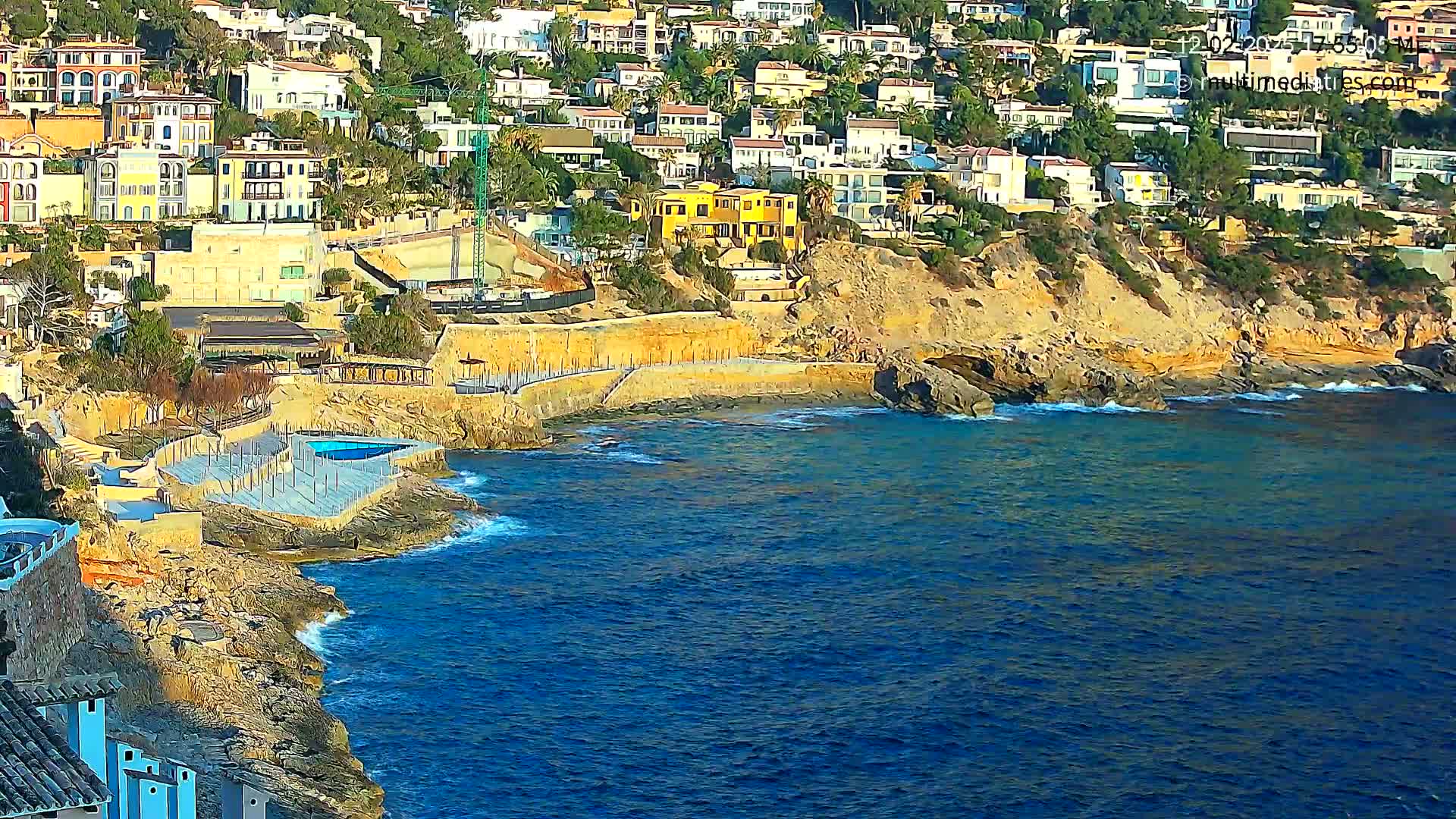 A rocky coastline with a small swimming pool and many houses on a hillside overlooks a calm, blue sea.