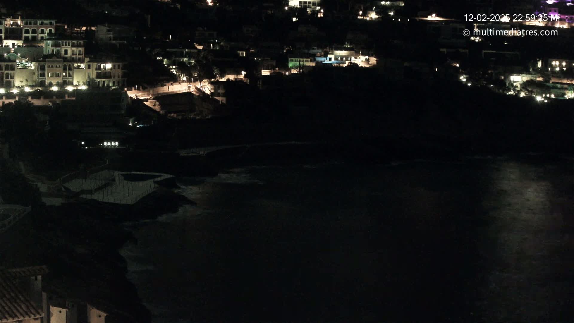 A nighttime coastal scene shows a dark body of water reflecting light, with a dark hillside featuring illuminated buildings above.