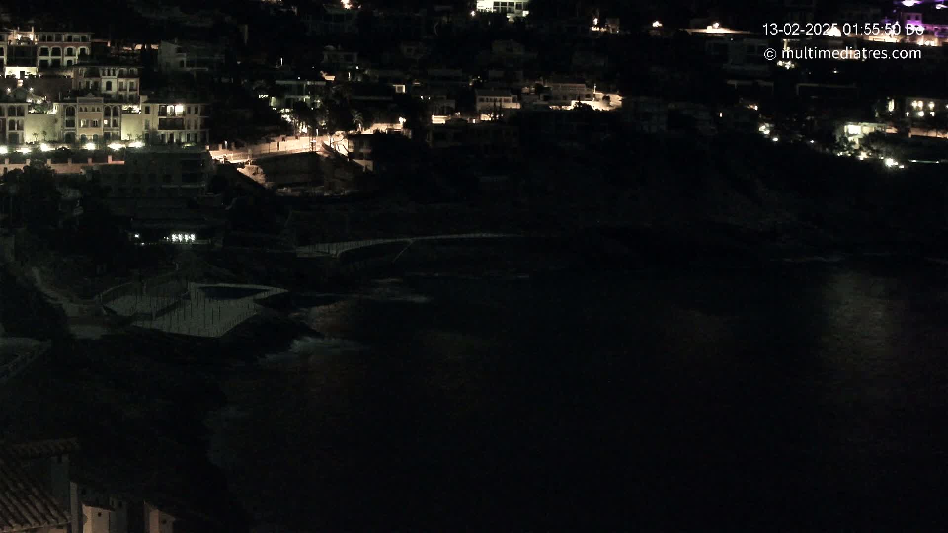 A nighttime view shows a dark body of water lapping at the shore below a collection of dimly lit buildings.