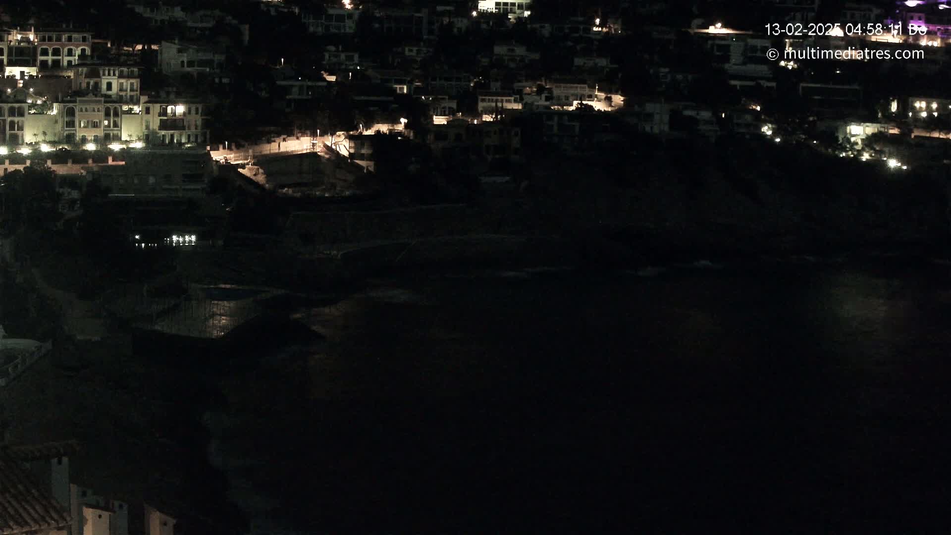 A nighttime view shows a dark body of water reflecting the lights of a hillside village.