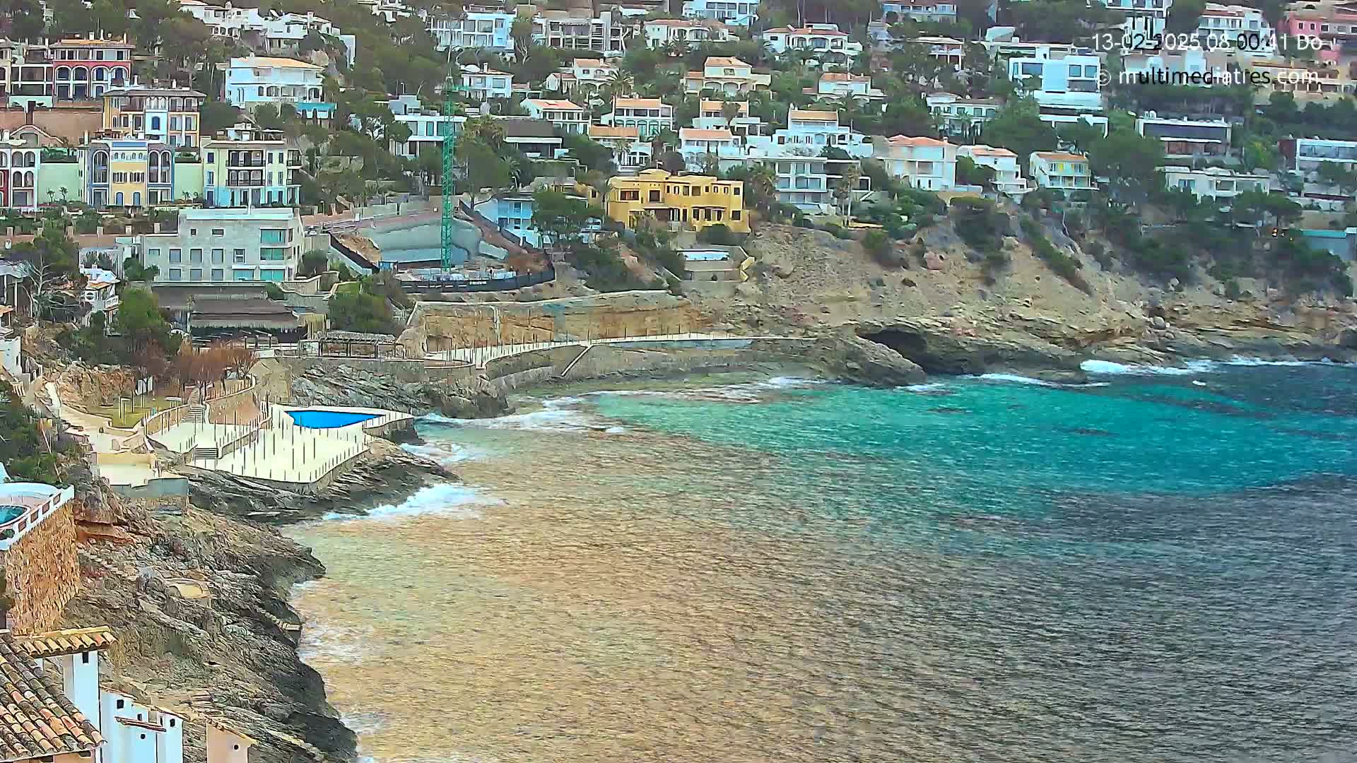 A rocky coastline with turquoise water, featuring a cluster of houses perched on a hillside, a small swimming pool, and some light wave action.