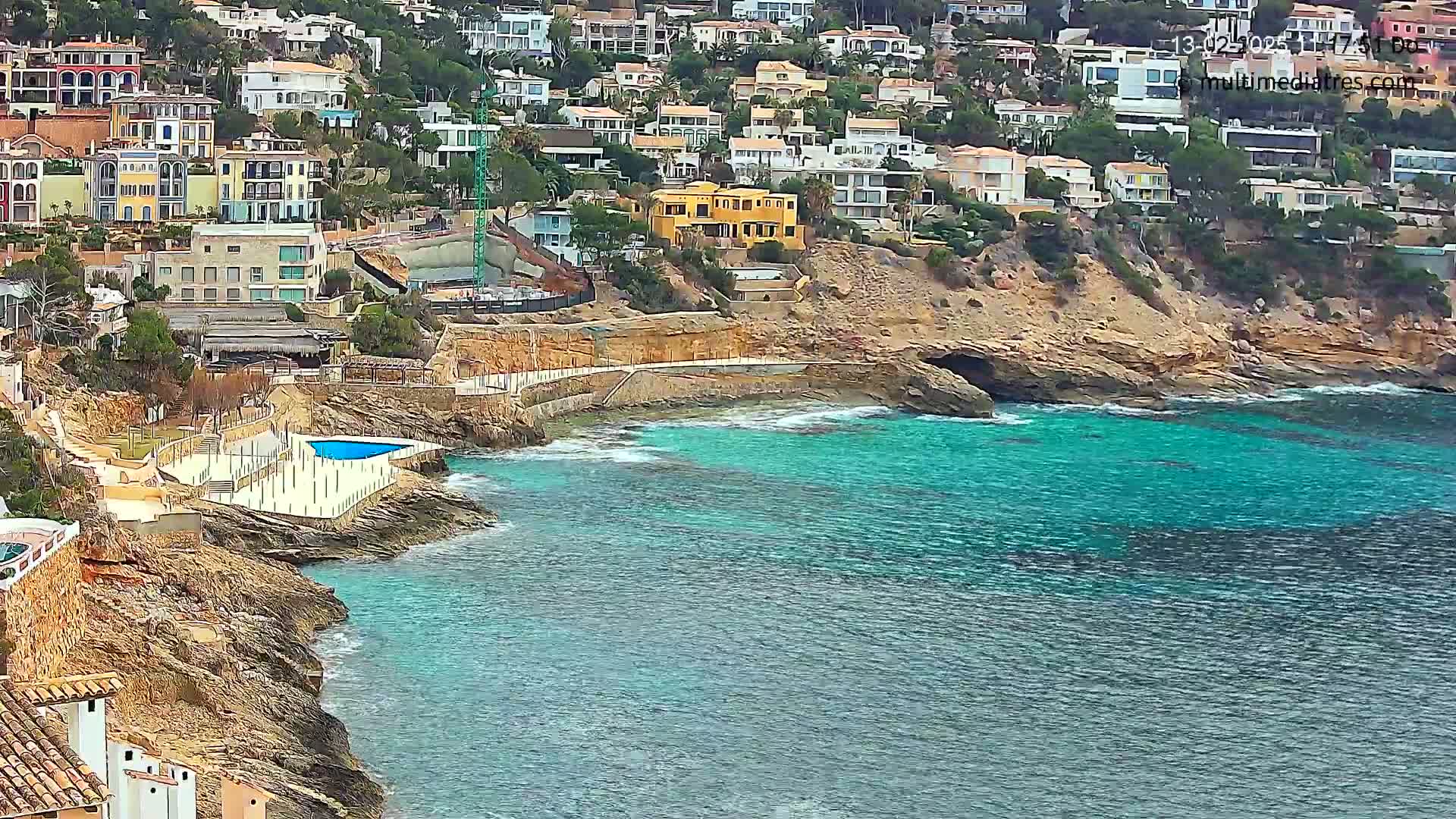 A rocky coastline with turquoise water features a cluster of houses on a hillside overlooking a small pool next to the sea.