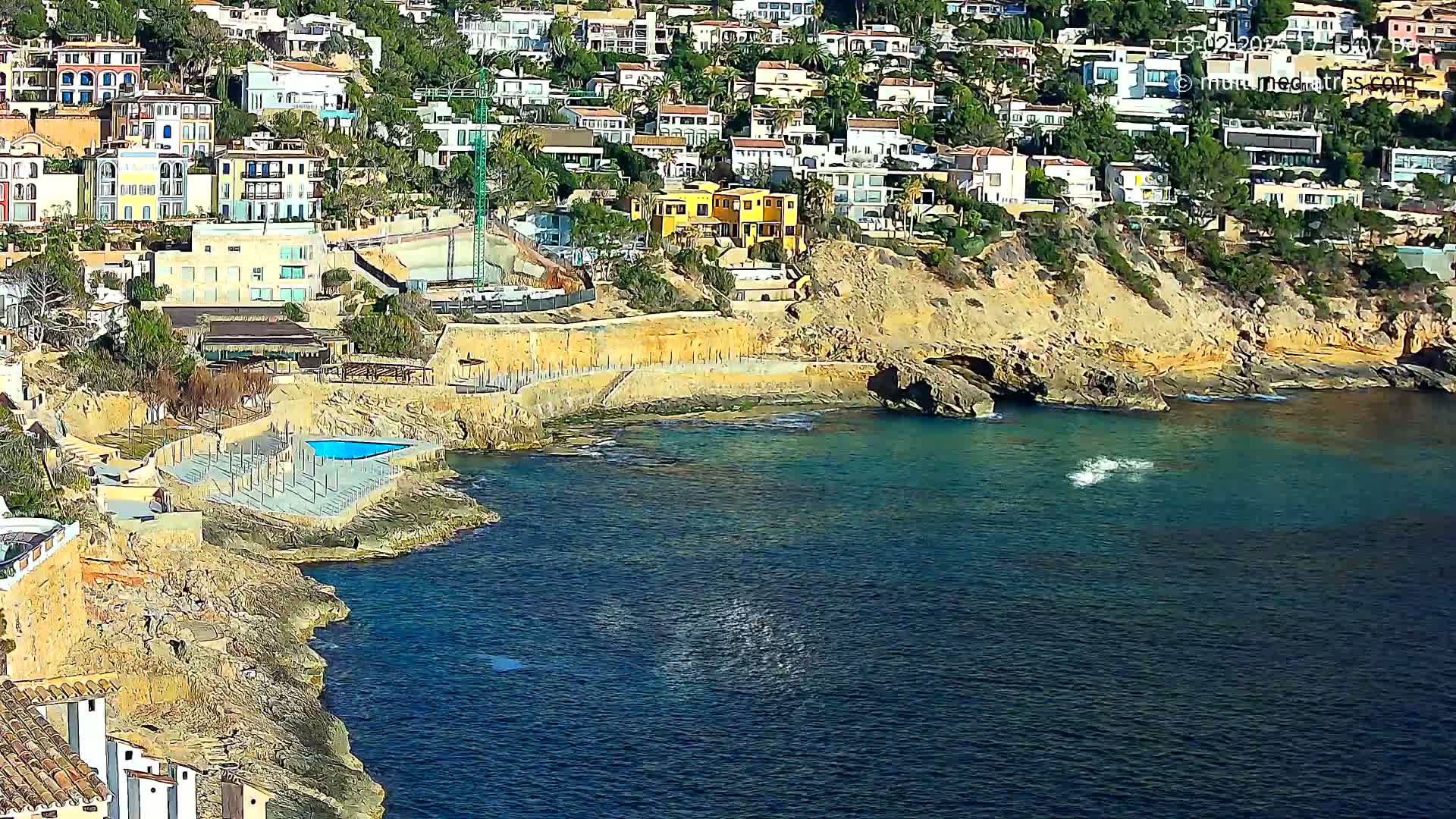 A coastal scene shows a cluster of buildings on a cliff overlooking a calm, blue sea with a small swimming pool next to the water.