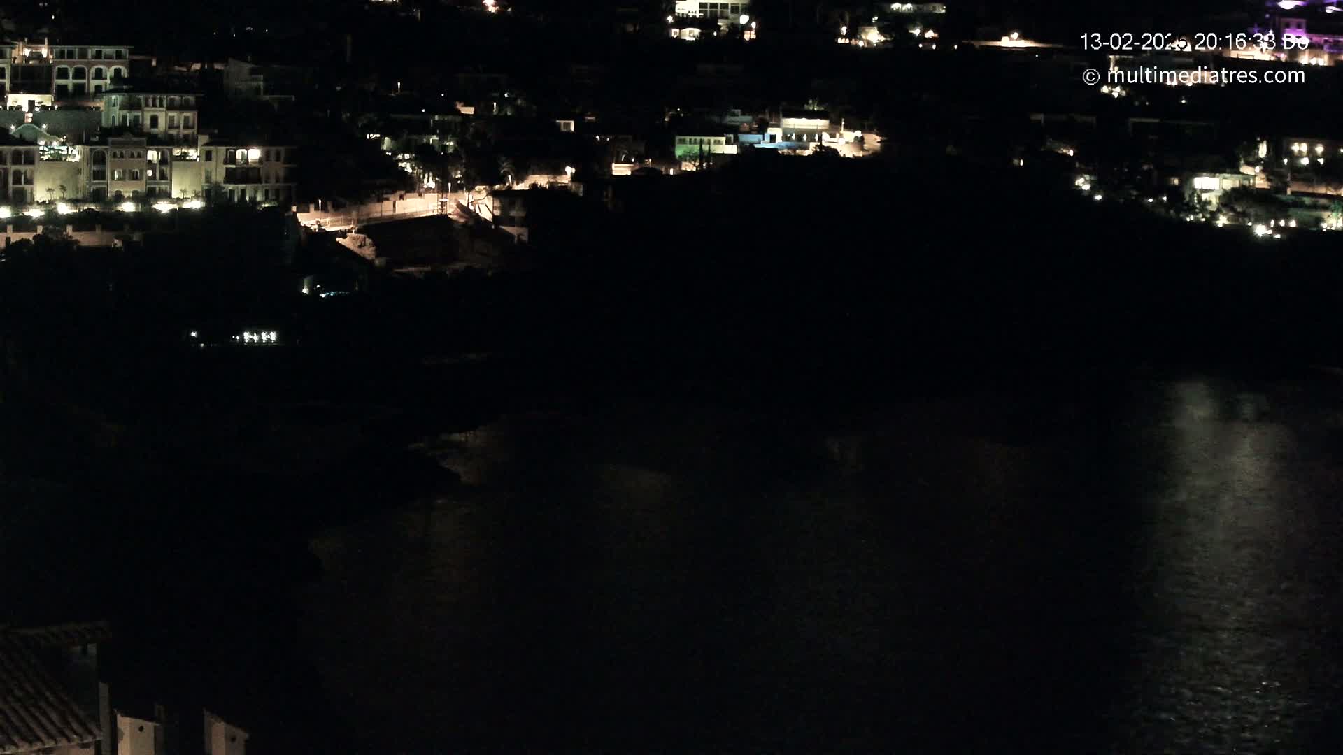 A nighttime view shows a collection of buildings nestled on a hillside overlooking dark water.