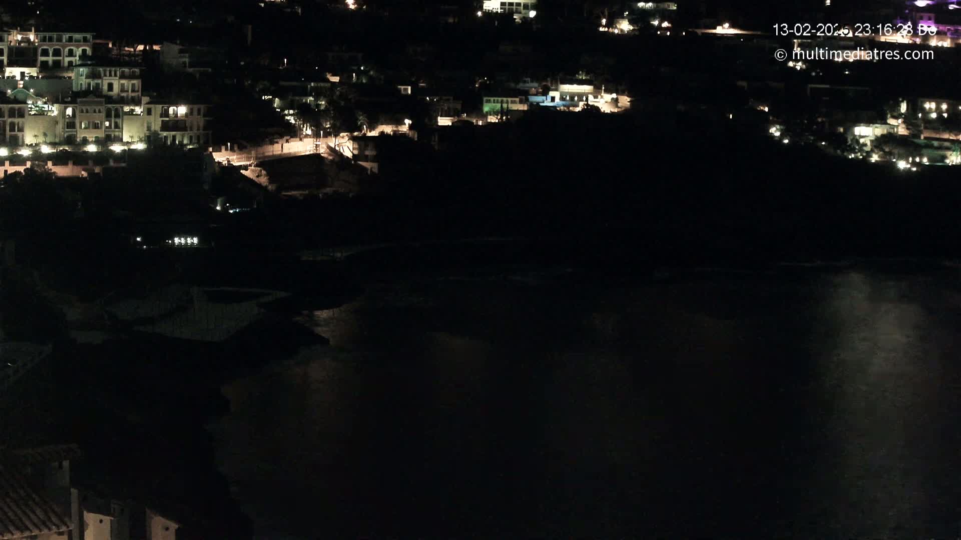 At night, a body of dark water reflects the lights of a hillside village.