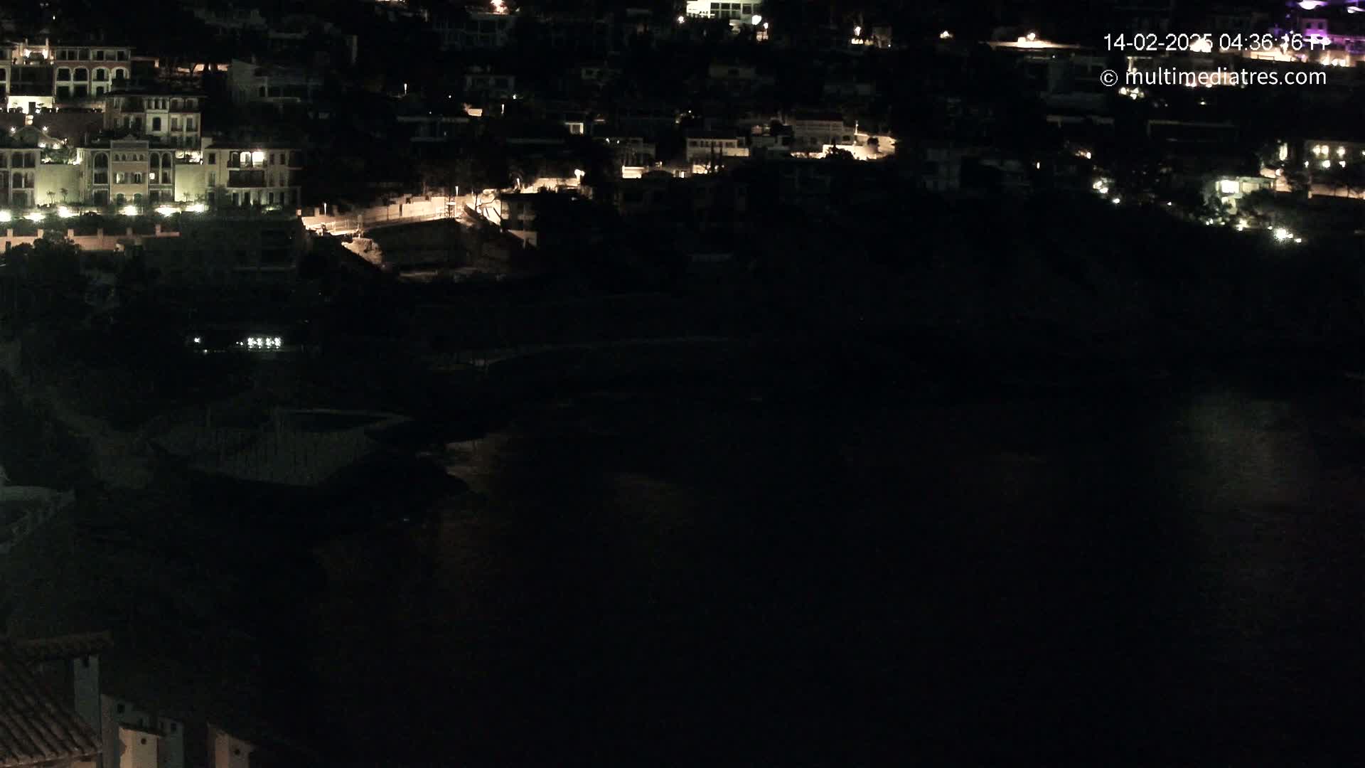 A nighttime, long shot of a coastal town with dimly lit buildings overlooking a dark body of water.