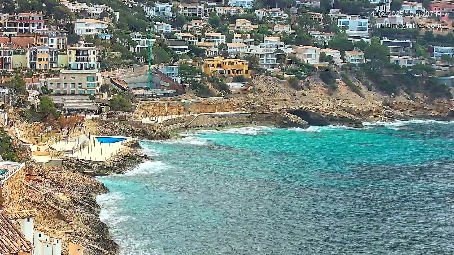 A rocky coastline with turquoise water features numerous houses built into a hillside, a small pool, and some light wave action.
