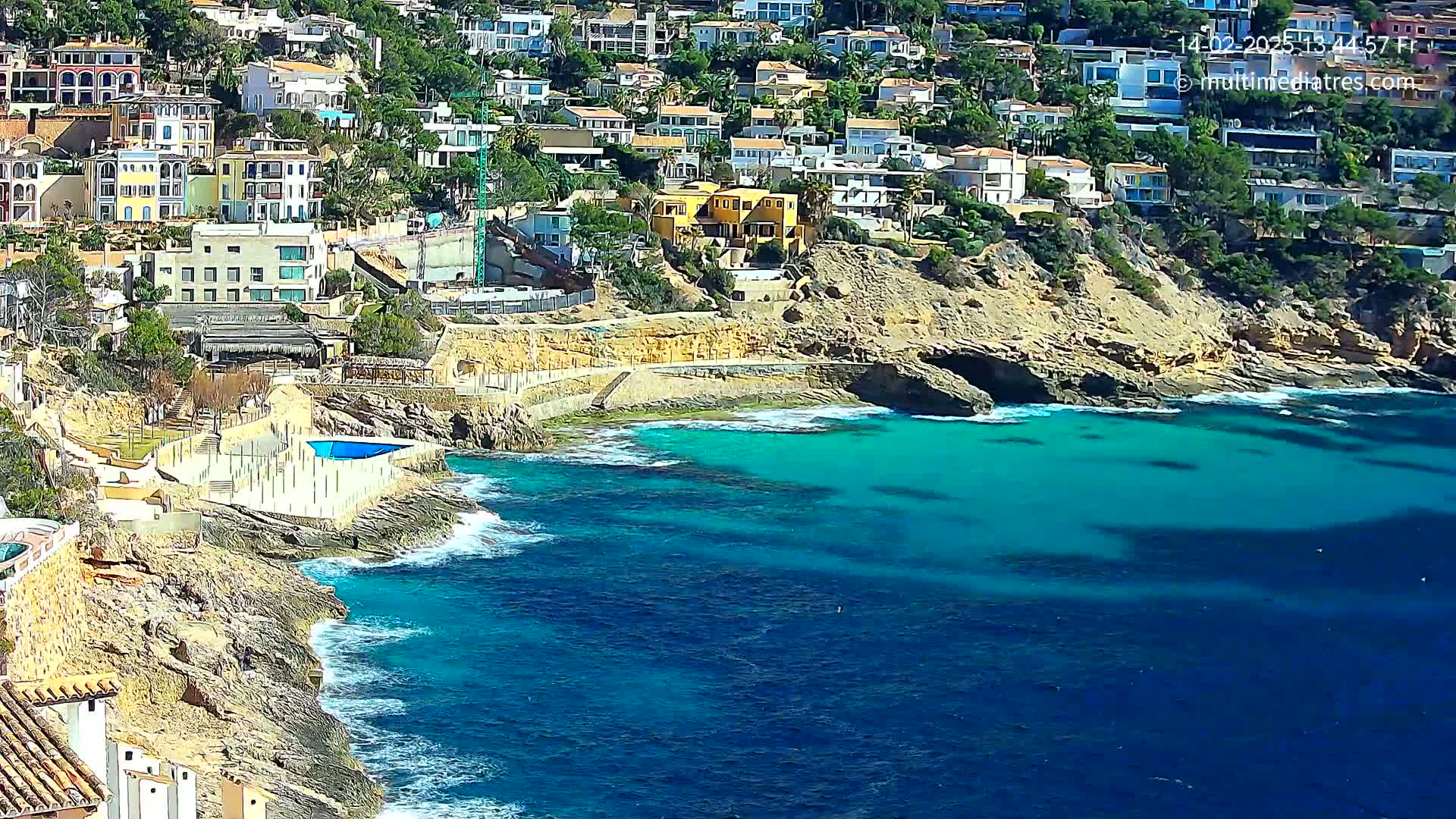 A rocky coastline with turquoise water features a cluster of houses built into the hillside, and a small swimming pool near the water's edge on a sunny day.
