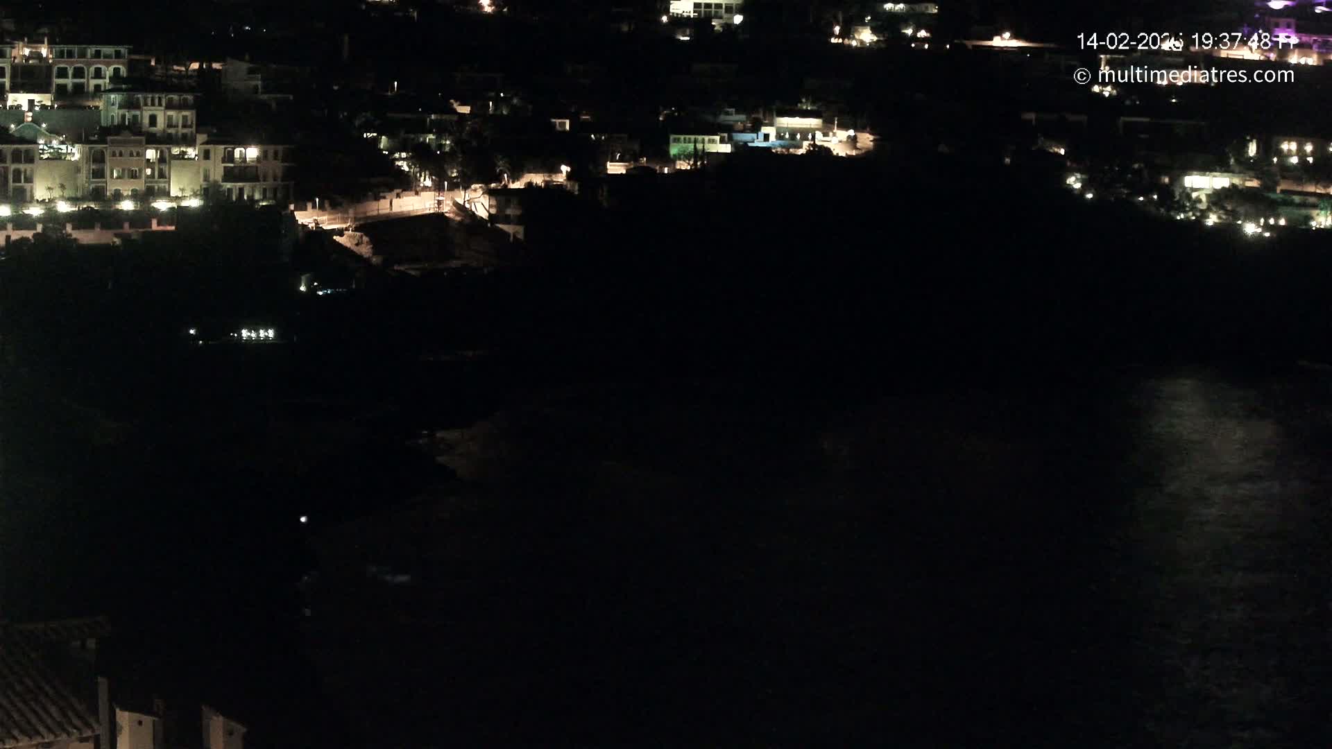 A nighttime aerial view shows a coastal town with illuminated buildings overlooking a dark body of water.
