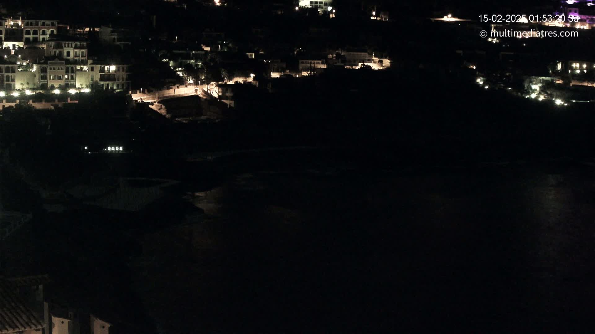 A nighttime, aerial view shows a dark body of water below a hillside of dimly lit buildings.