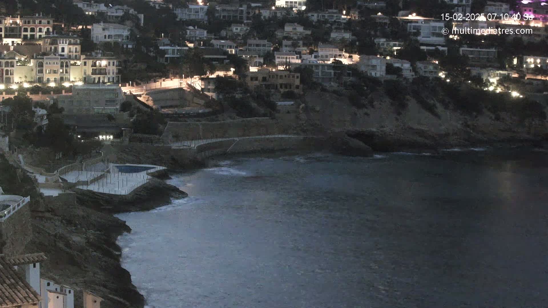A coastal town at twilight, with a small swimming pool situated on a rocky cliff overlooking a calm sea.