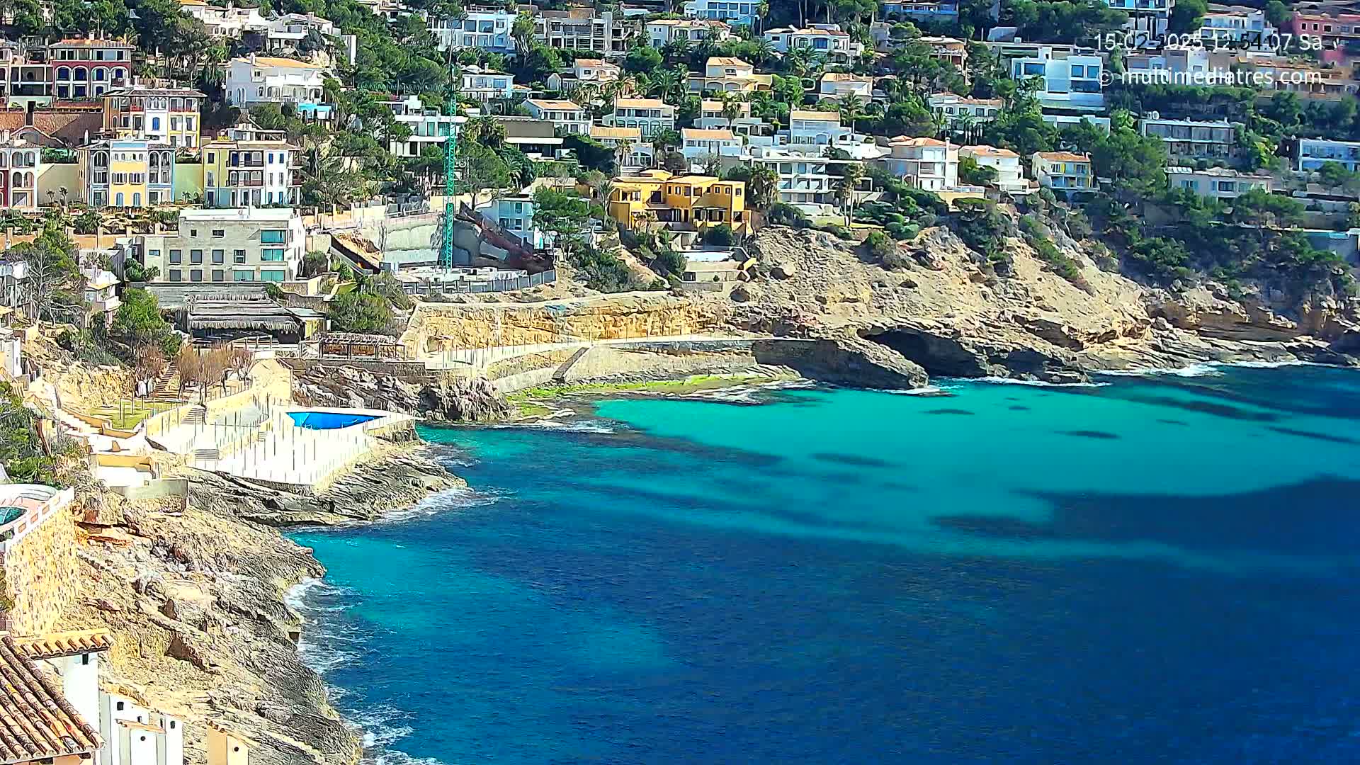 A rocky coast with bright turquoise water features a collection of white and yellow buildings on a hillside.