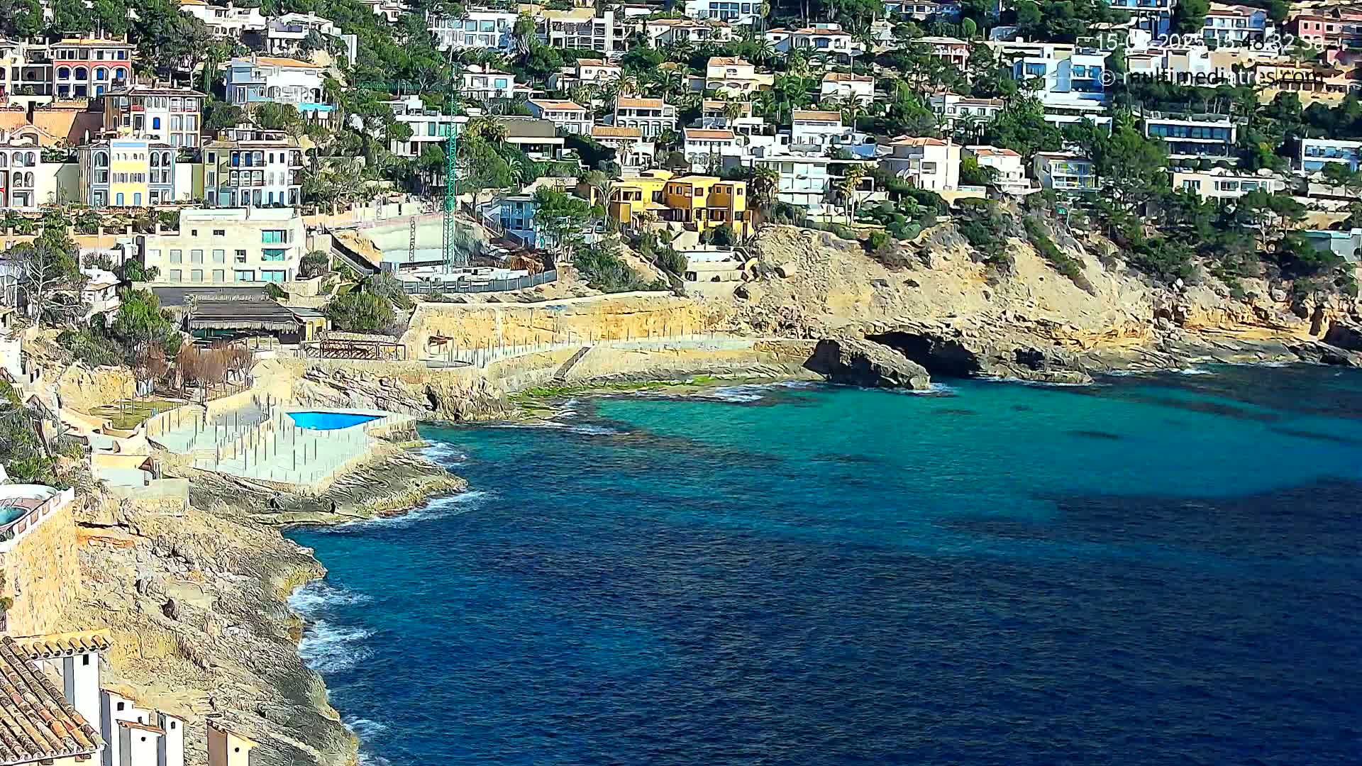 A rocky coastline with turquoise water features a cluster of houses perched on a hillside, a small pool, and some ongoing construction.