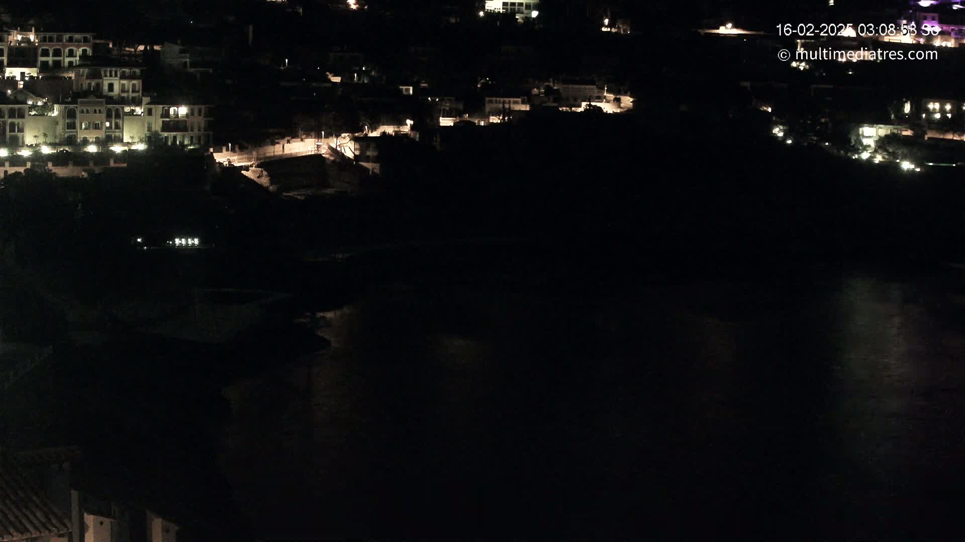 A nighttime, long shot shows a dark body of water with a hillside of dimly lit buildings in the background.