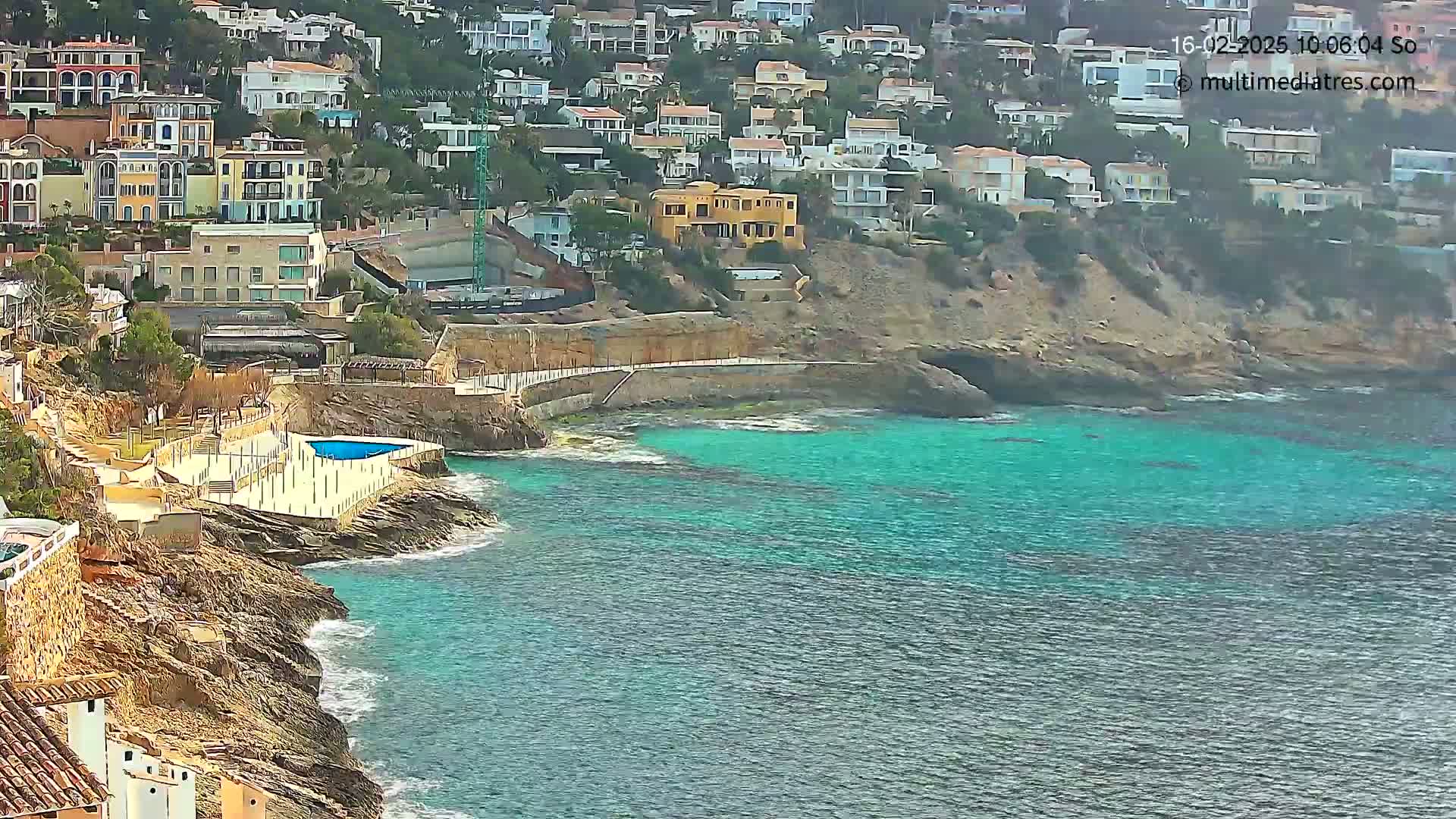 A rocky coastline with turquoise water features a small pool and many houses built on a hillside, under a mostly clear sky.