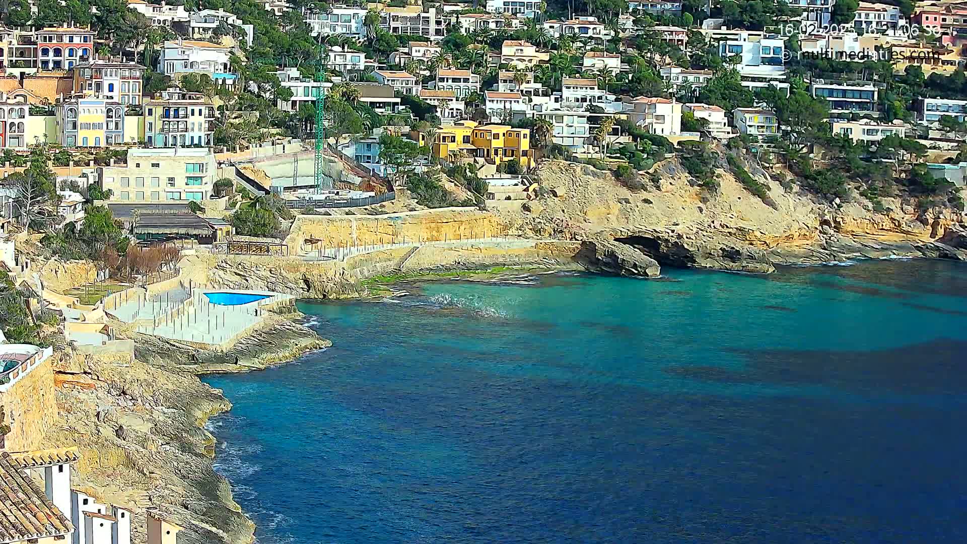 A rocky coastline with turquoise water features many houses built into a hillside, and a small swimming pool is visible near the water's edge.