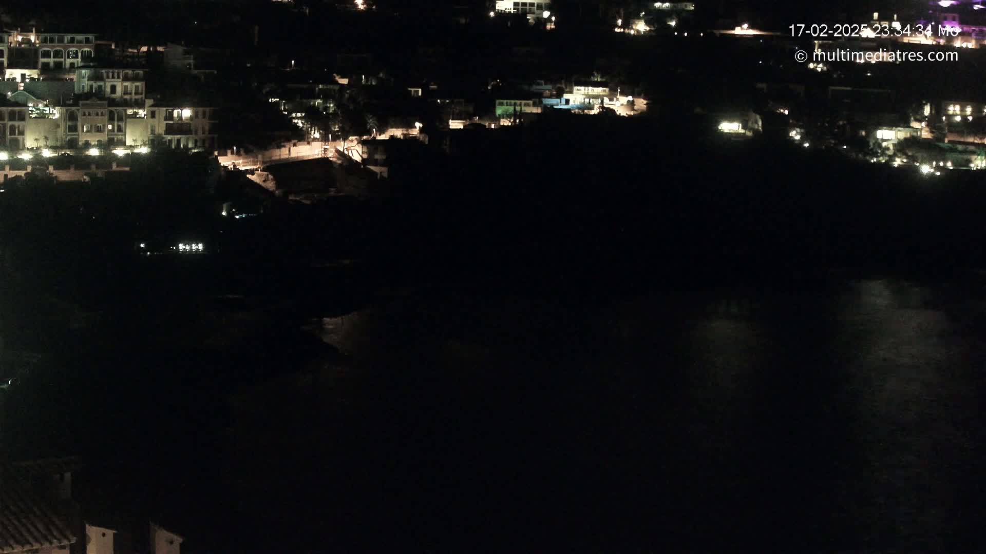 A nighttime, aerial view shows a coastal town with dimly lit buildings and a dark body of water.
