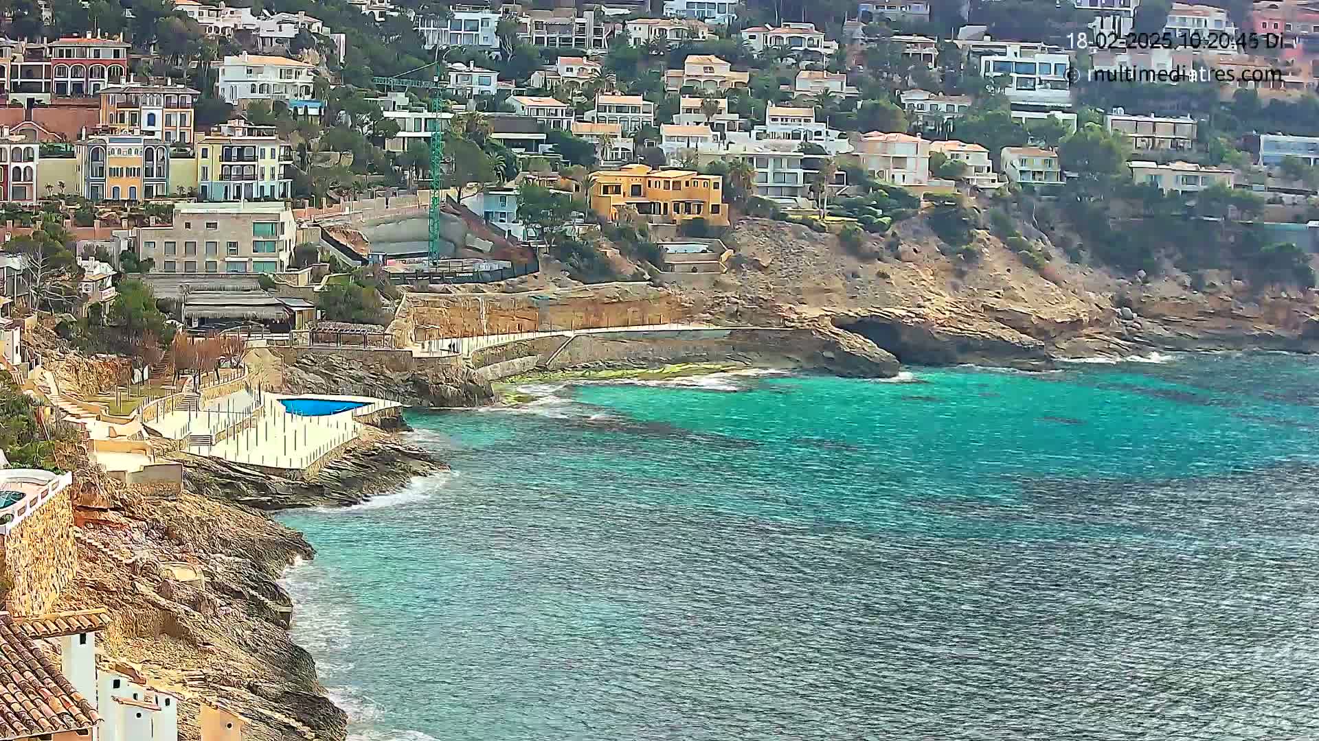 A coastal town with numerous houses built on a hillside overlooking a teal-colored sea on a partly cloudy day.