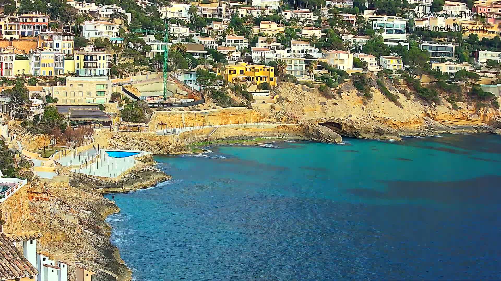 A rocky coastline with turquoise water features a small swimming pool next to many houses built into a hillside.