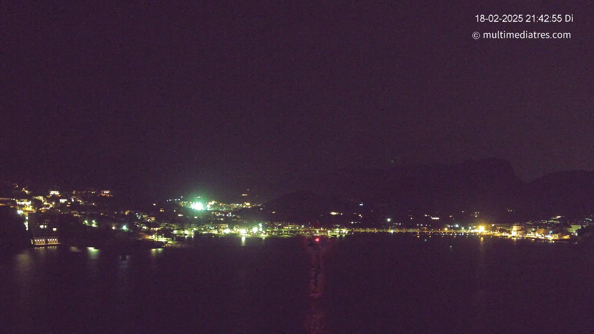 A nighttime view of a town situated on a hill overlooking a dark body of water under a clear, dark sky.