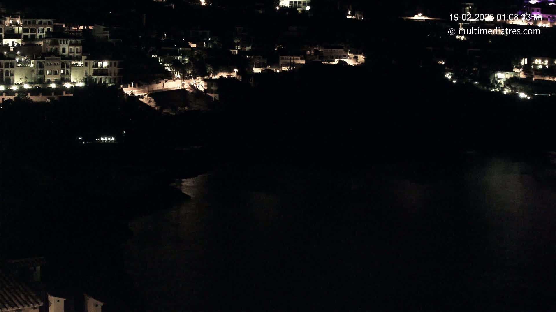 A nighttime aerial view shows a dark body of water next to a hillside village with sparsely lit buildings.