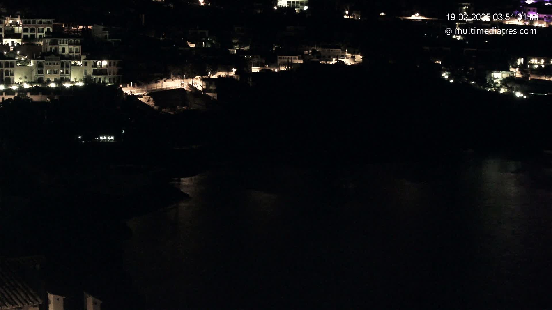A nighttime, high-angle view shows a dark body of water next to a hillside of dimly lit buildings.