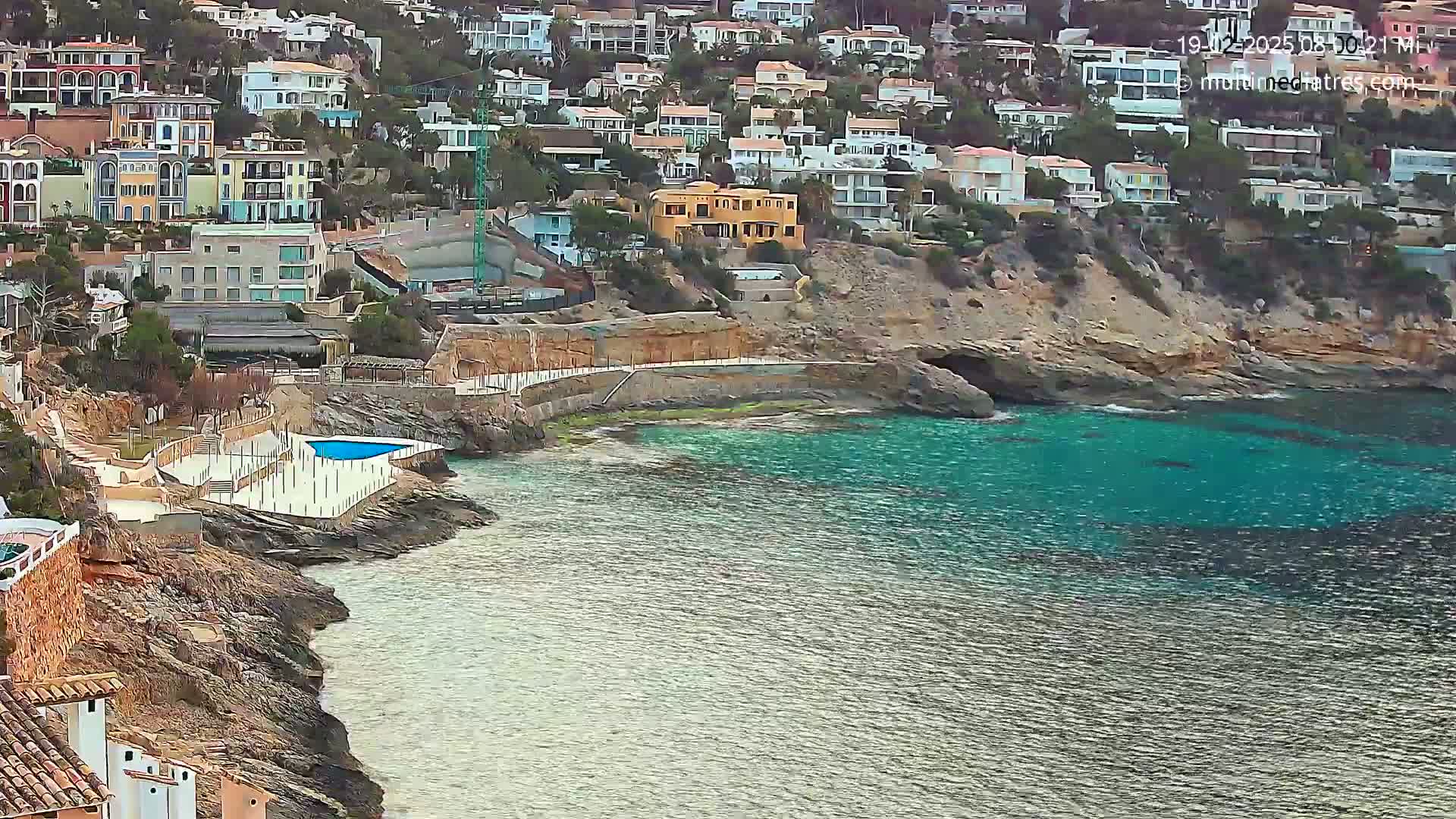 A rocky coastline with turquoise water features a small pool and many houses built into the hillside.