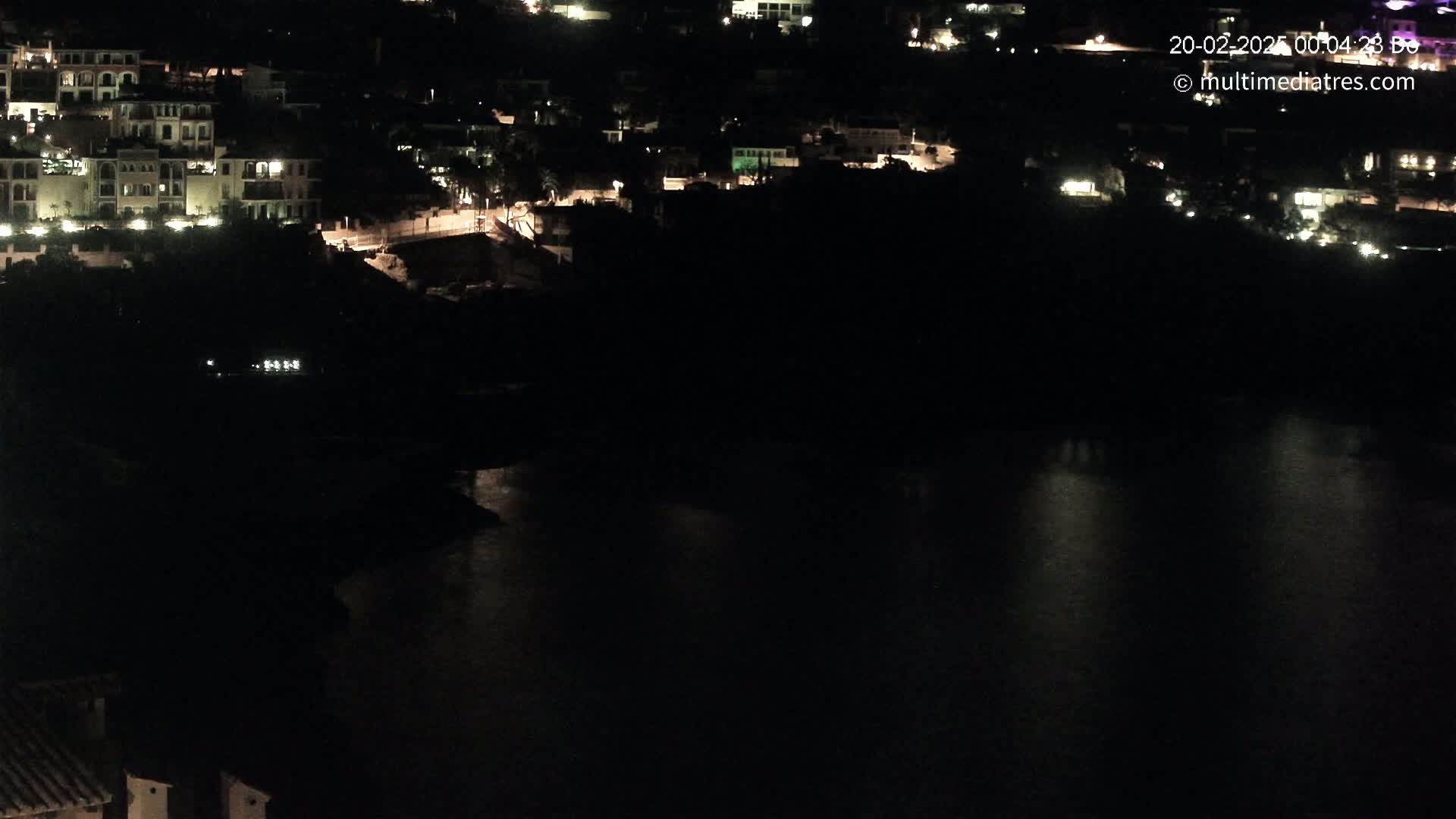 A nighttime view of a coastal town with buildings lit up, overlooking a dark body of water.
