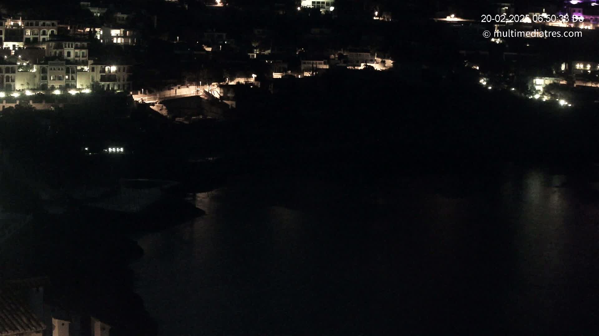 A nighttime, aerial view shows a dark body of water reflecting scattered lights from buildings nestled on a hillside.