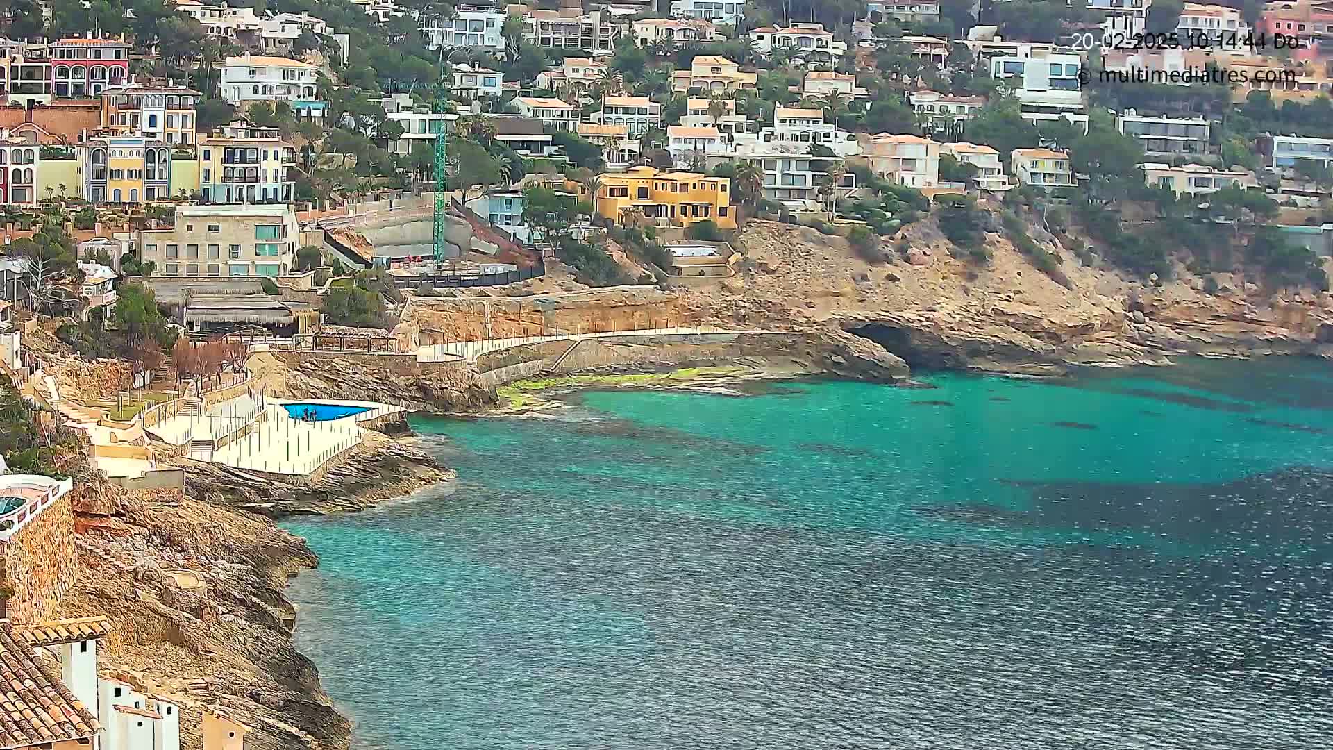 A rocky coastline with turquoise water features a small pool and numerous houses built into a hillside.