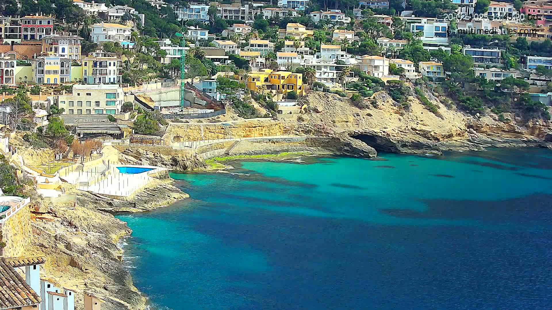 A coastal scene shows a rocky hillside dotted with houses overlooking a calm, turquoise sea, under sunny skies.